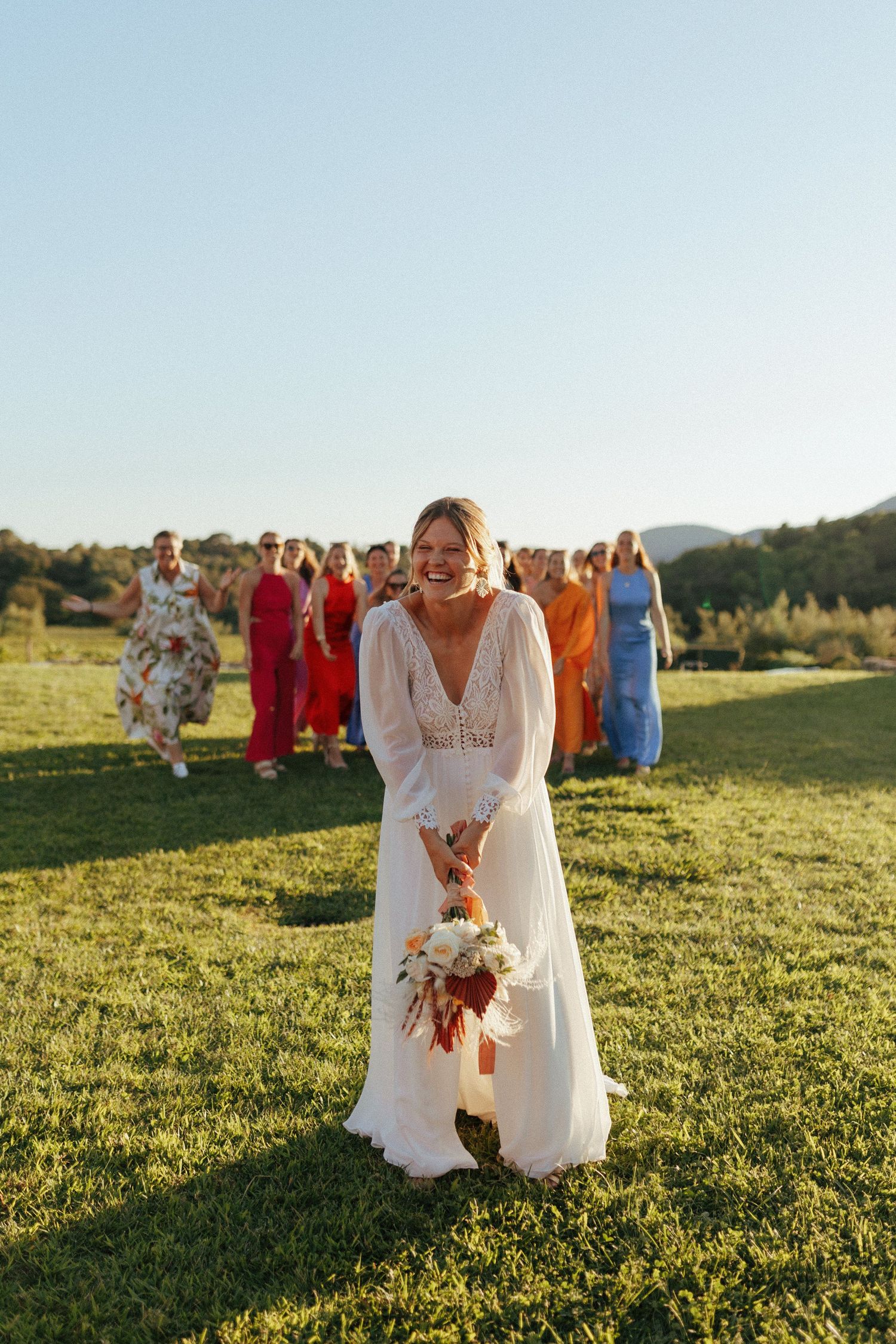 A bride in a flowing white dress stands in a sunlit meadow with wedding guests in colorful dresses behind her.