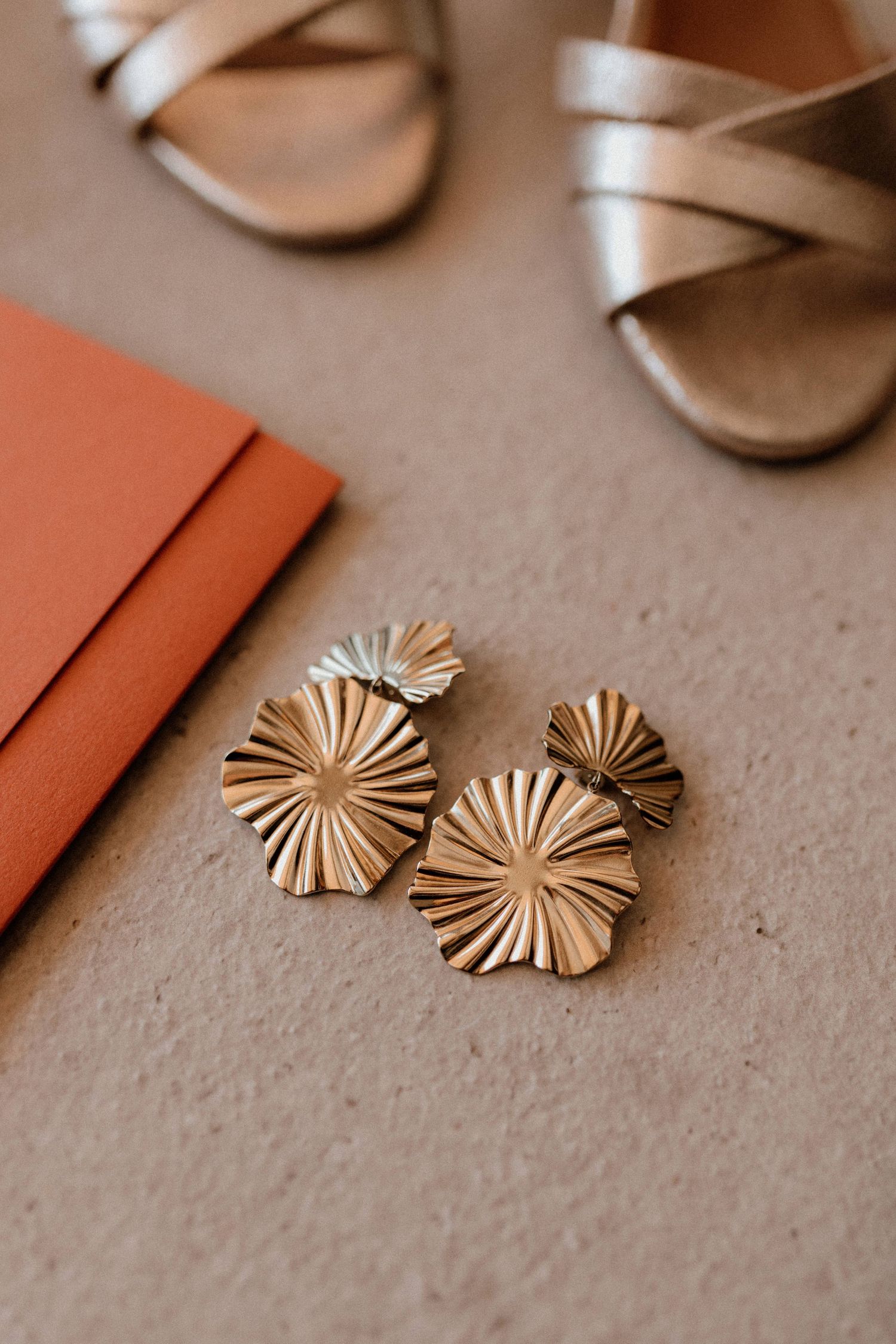 Close-up of gold sunburst statement earrings next to coral wedding stationery and silver sandal.