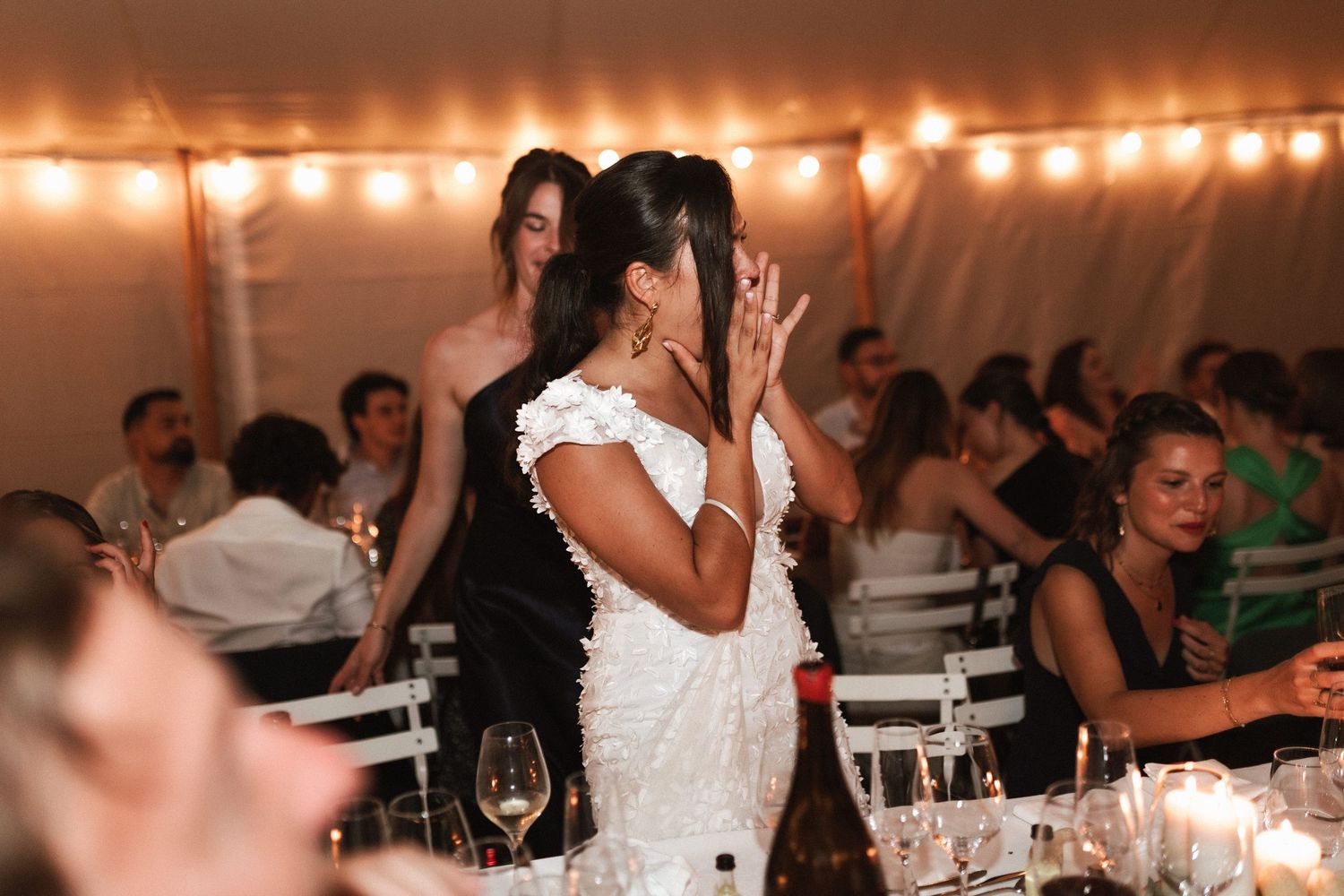 Wedding reception guests celebrate under string lights in a white tent as someone in a white dress stands at the dinner table.