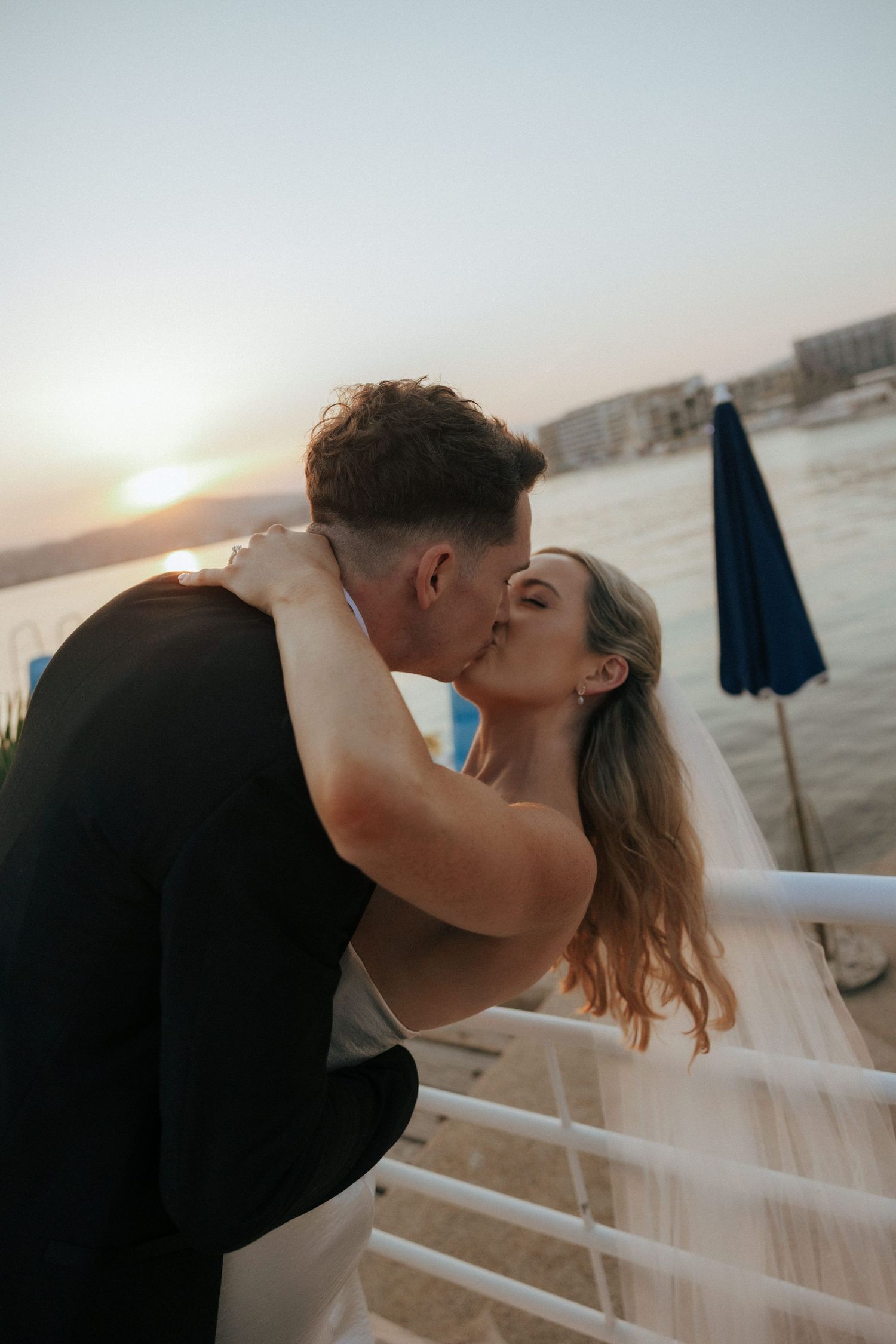 A romantic sunset moment as a newly married couple share a kiss on a waterfront terrace with a veil flowing in the breeze.