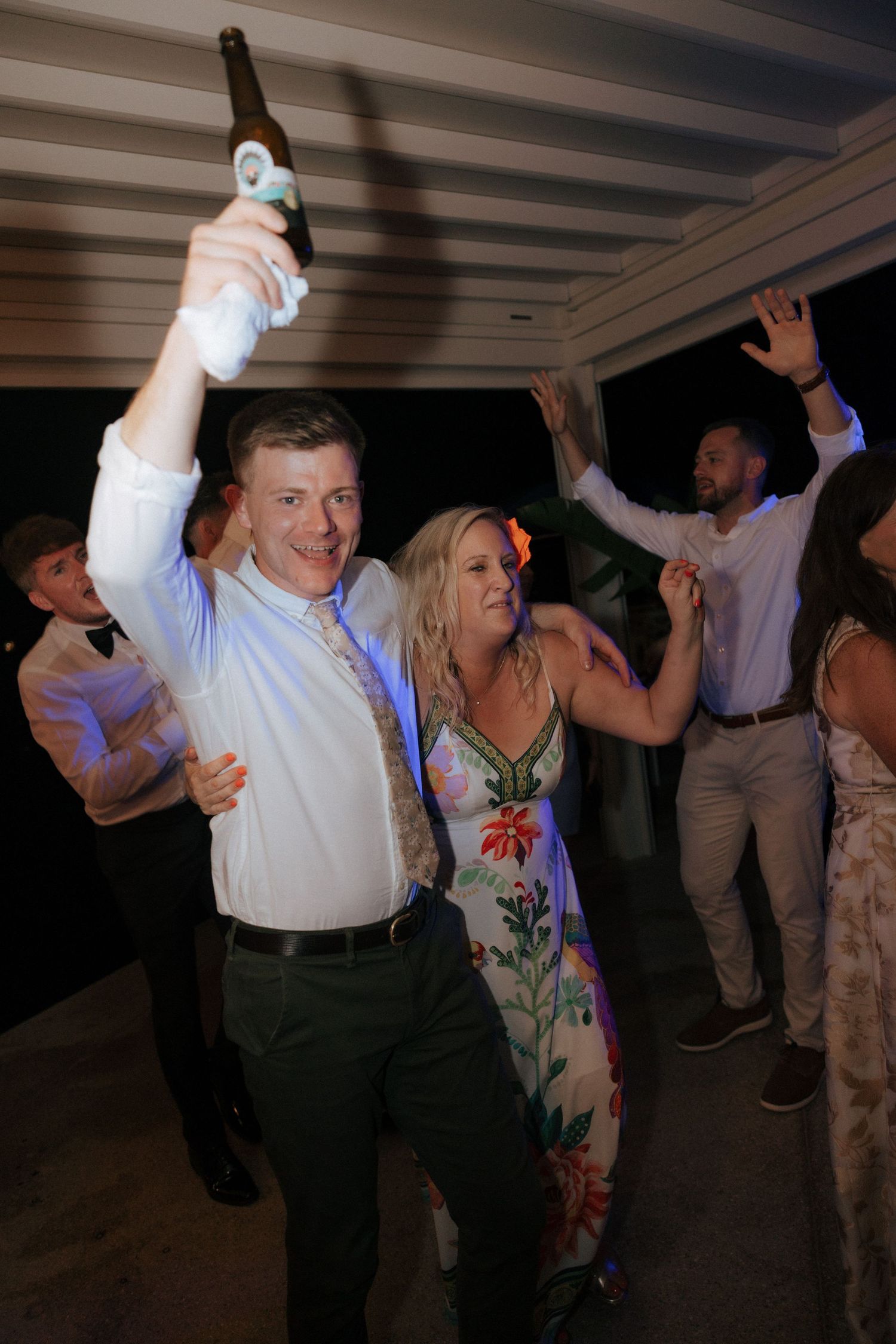 Wedding guests dance and celebrate on the dance floor with one person holding up a beer bottle at an indoor reception.