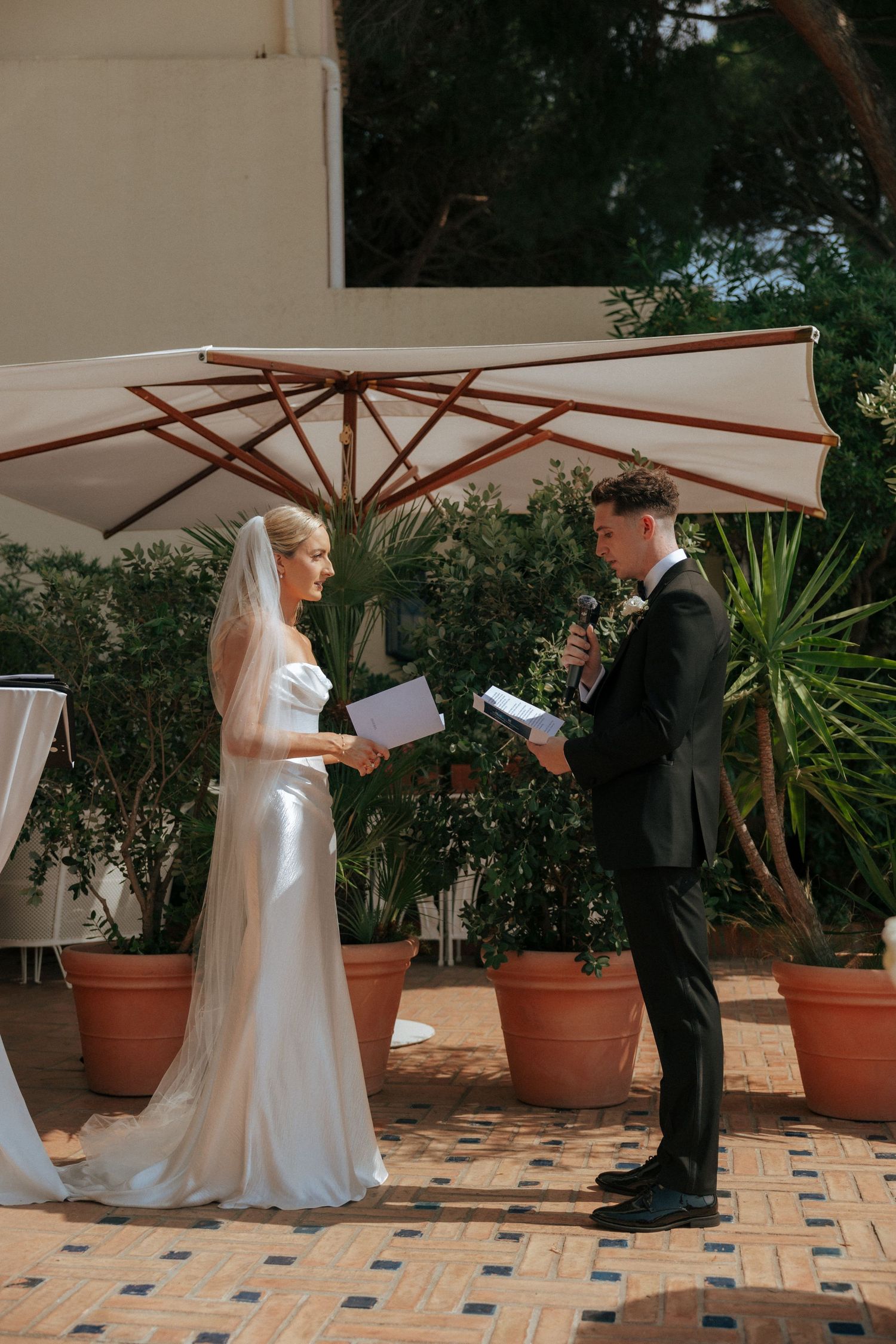 A wedding ceremony on a tiled patio with potted plants and a white umbrella overhead.