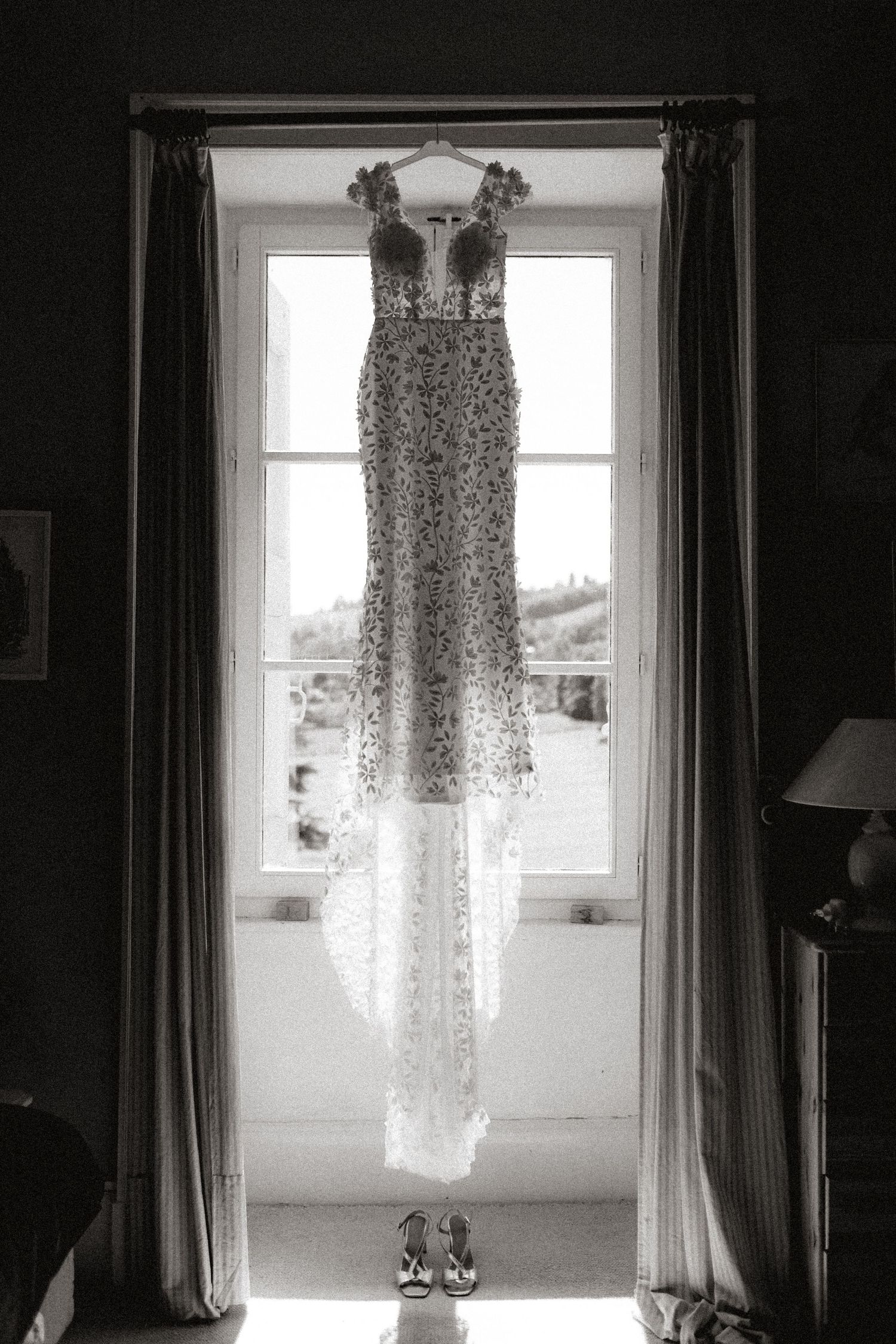 Lace wedding dress hangs in window frame creating ethereal silhouette.