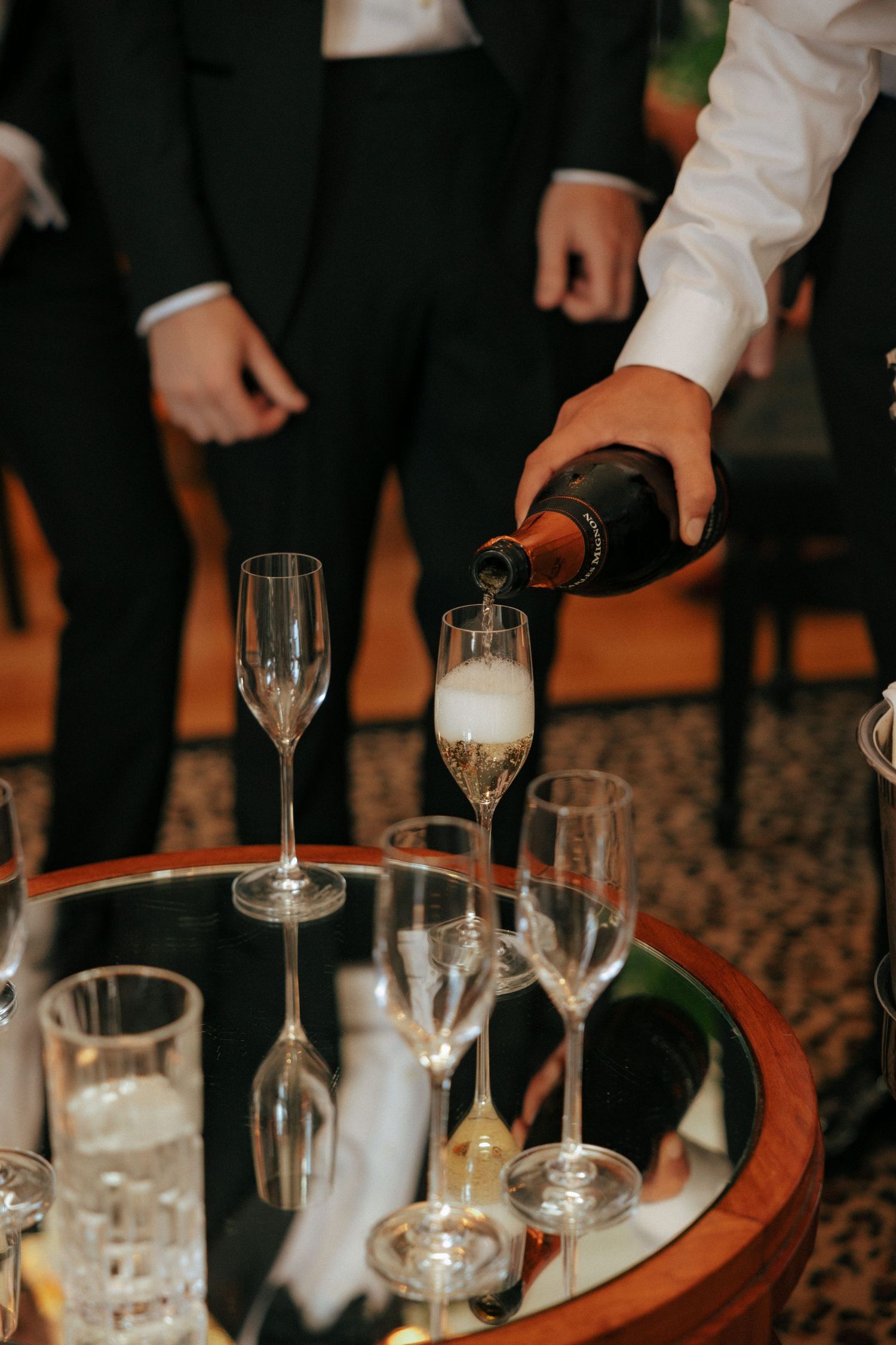 Close-up view of hands pouring drinks into champagne glasses on a wooden bar surface.