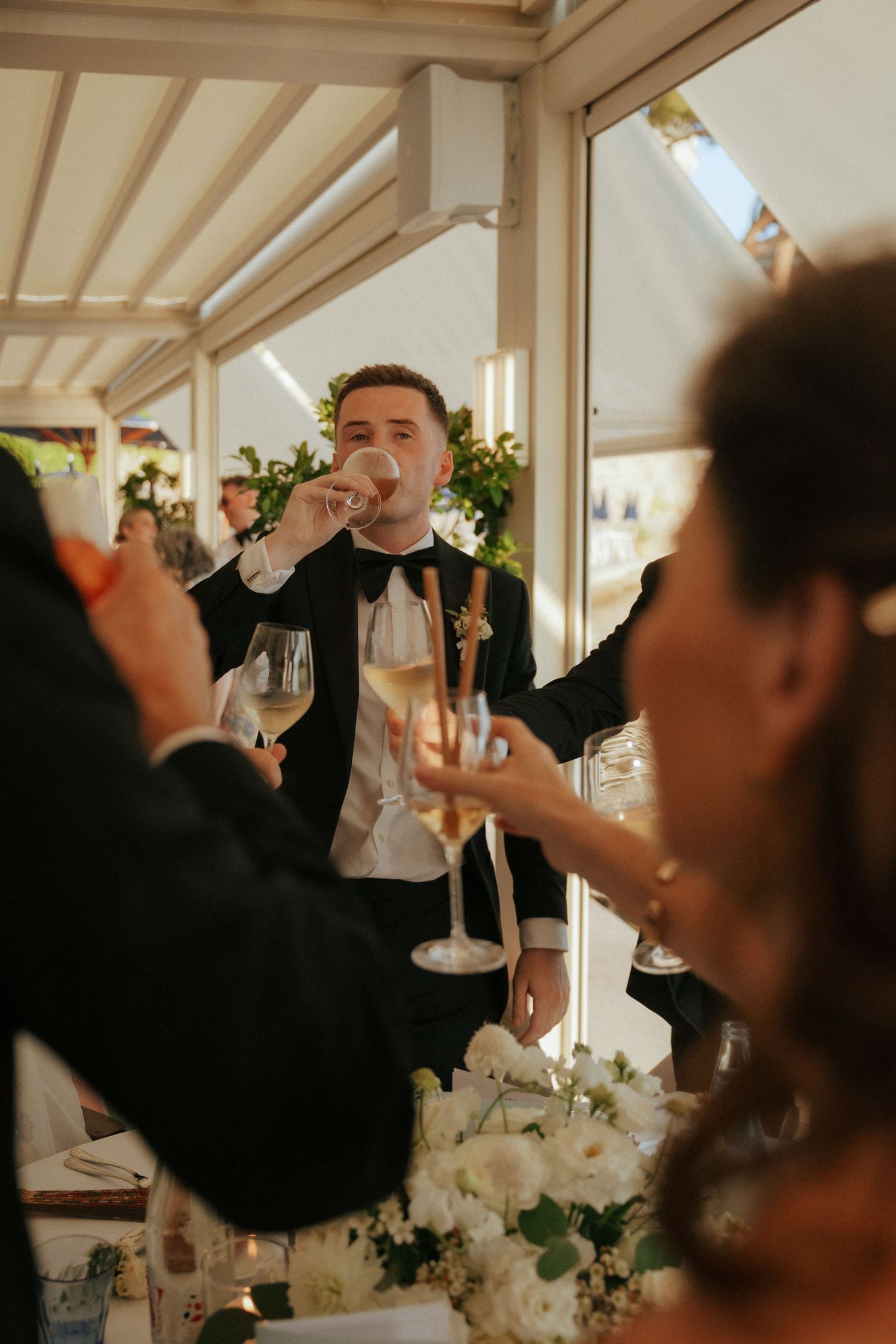 Elegant wedding guest in black tuxedo making a toast at a reception with floral centerpieces.