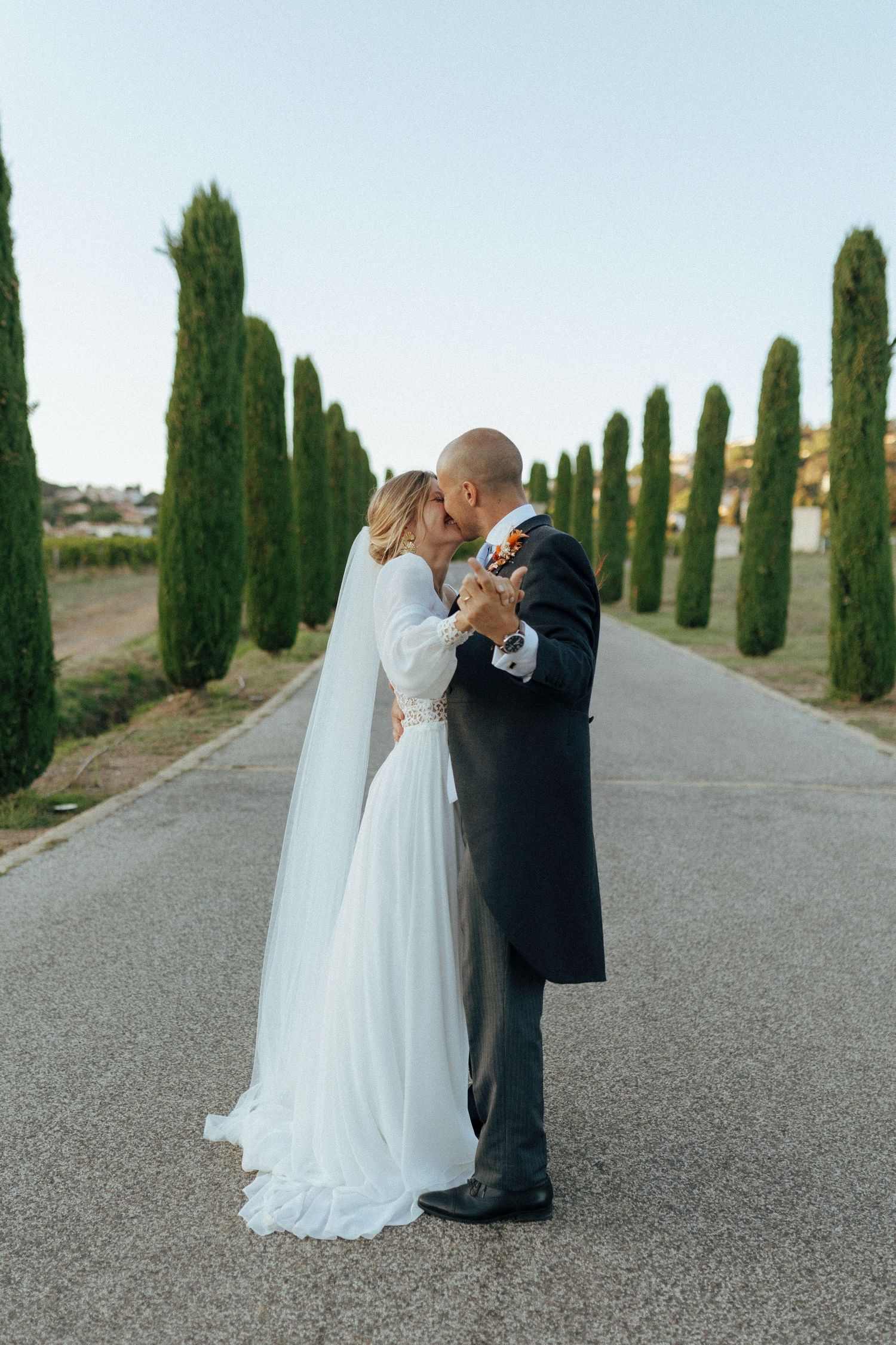Wedding couple sharing intimate moment on tree-lined road with cypress trees in Mediterranean setting.