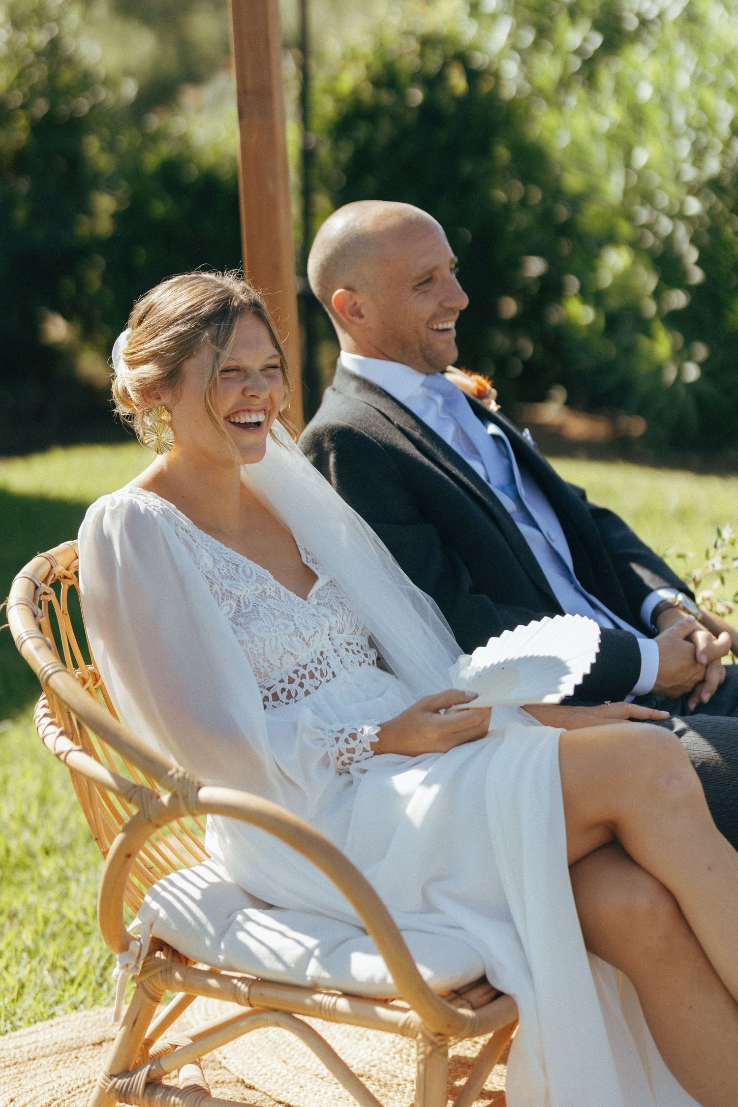Wedding couple sharing a joyful moment while seated in rattan chairs outdoors.