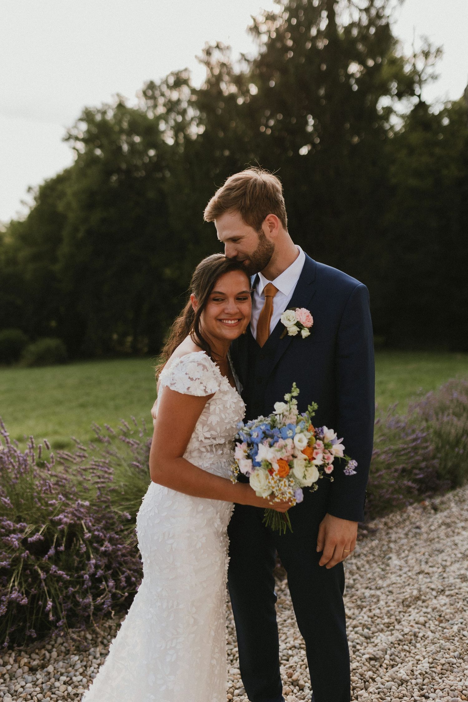 A wedding couple poses together outdoors with a beautiful bouquet of mixed flowers on a gravel path.
