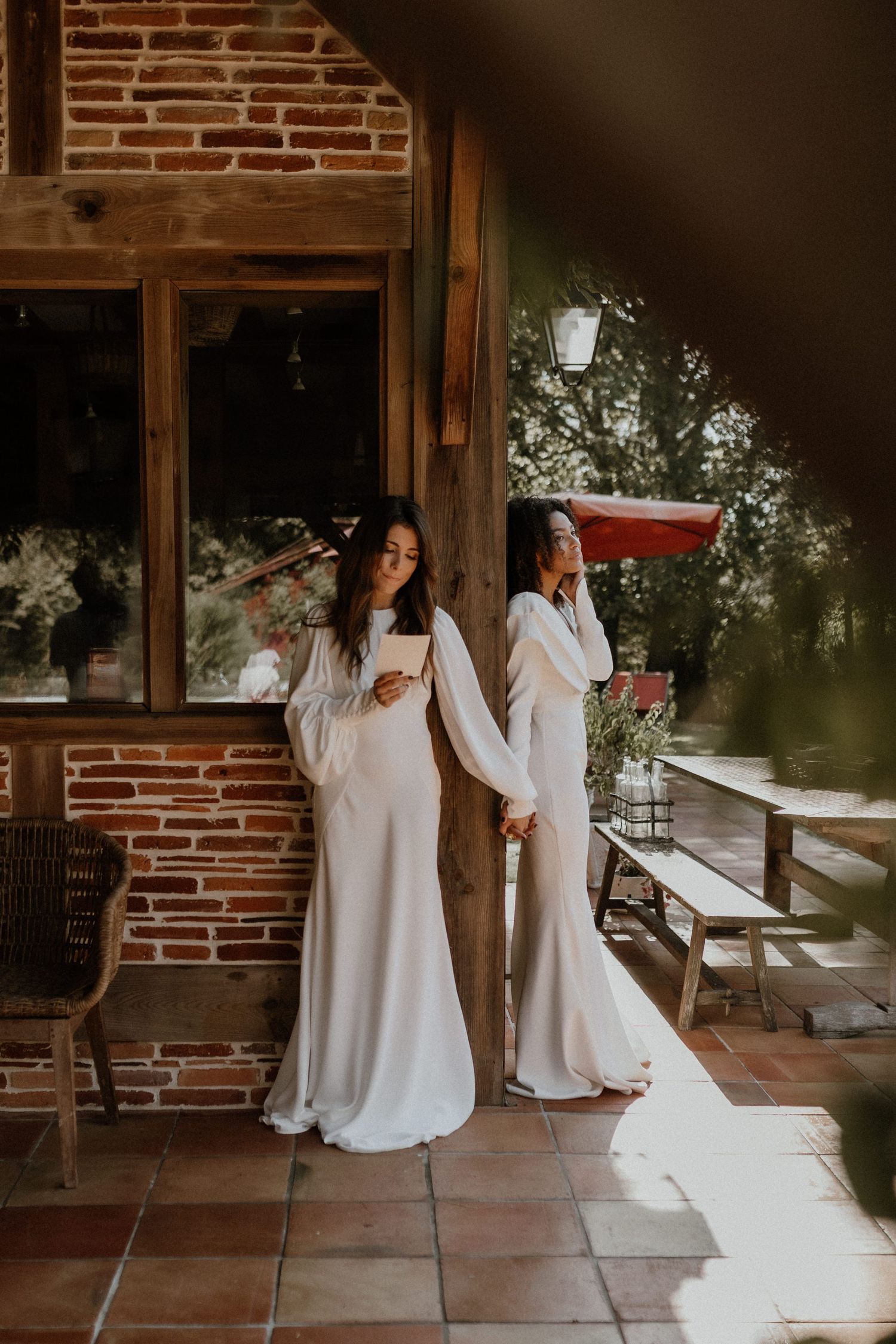 Two women in flowing white gowns stand in front of a rustic brick wall with a white lantern and sunlit patio.