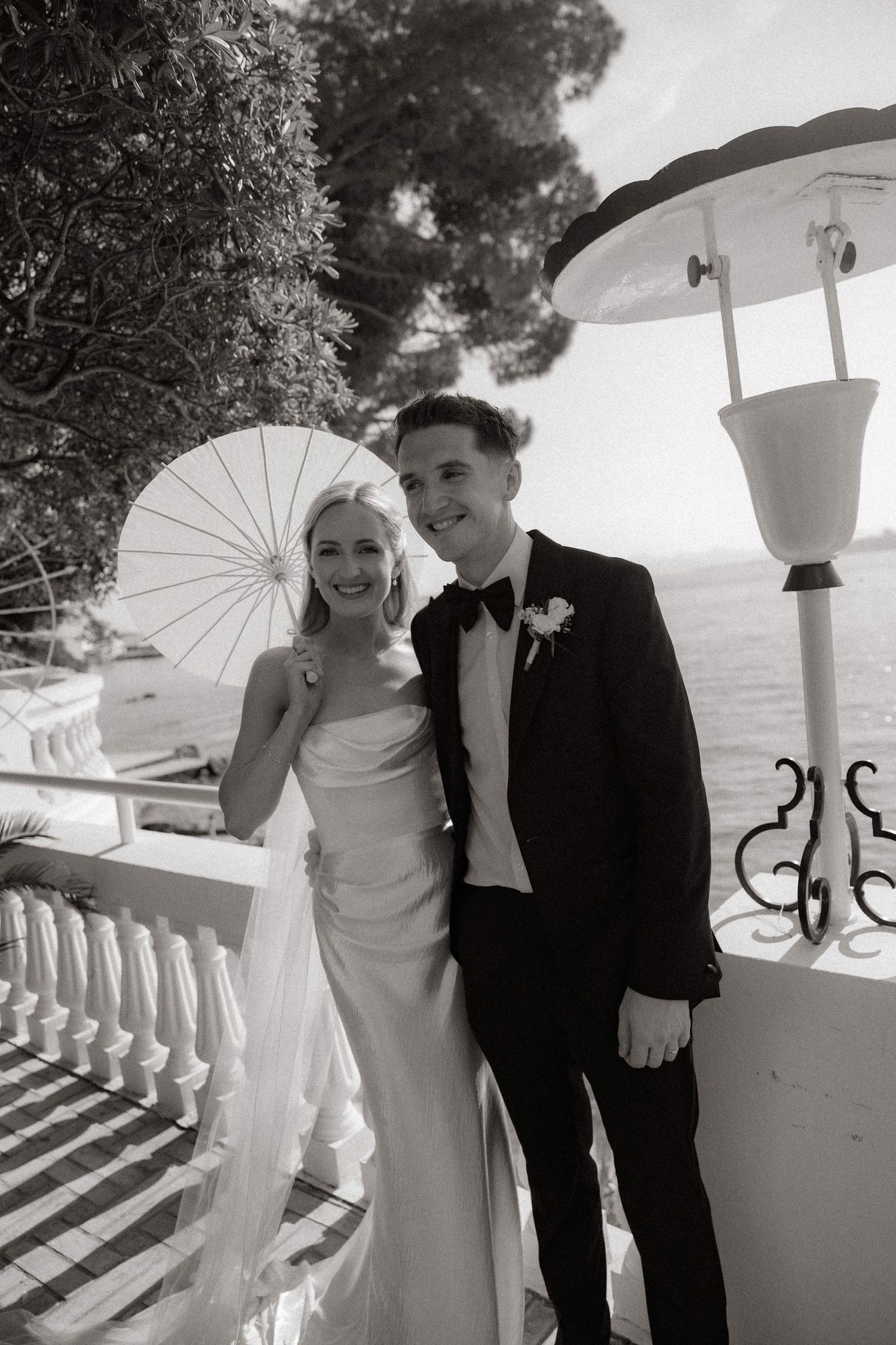A classic black and white wedding photo features a couple with a parasol on an elegant balcony overlooking the ocean.