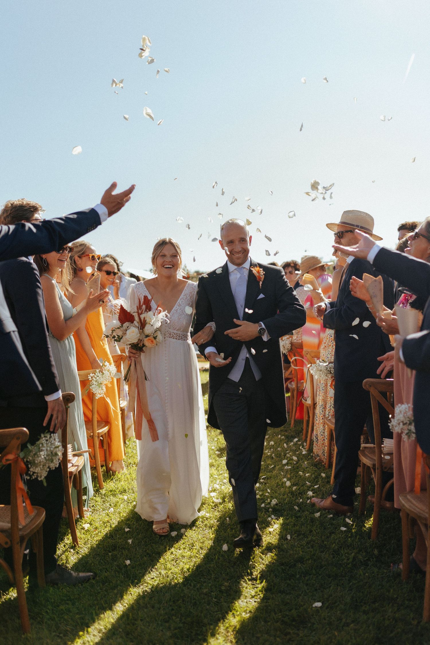 Wedding guests throw confetti as a couple walks down the aisle at a sunny outdoor ceremony.