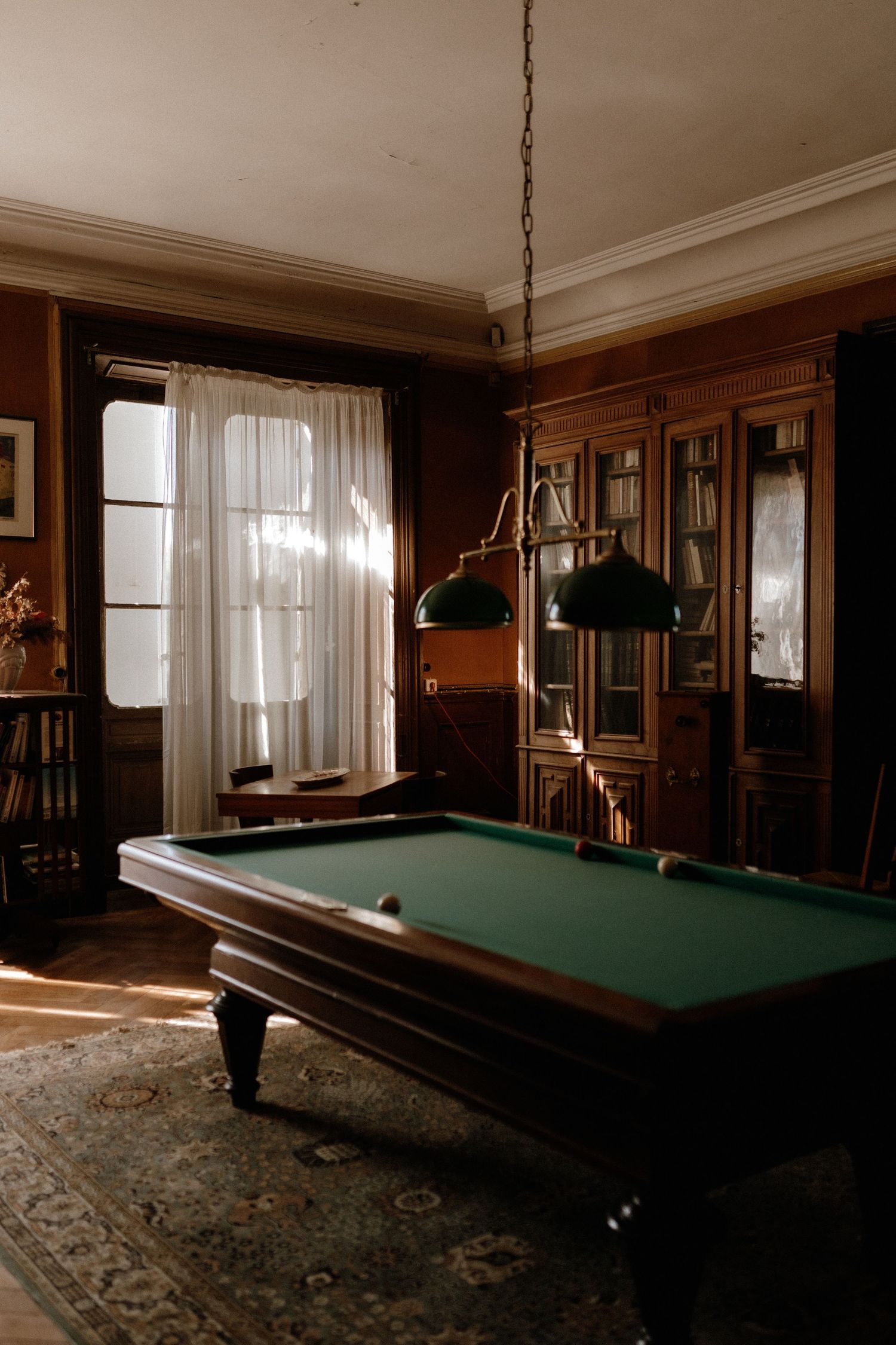 A moody billiards room with wood paneling features a pool table, vintage lighting, and large windows with sheer curtains.
