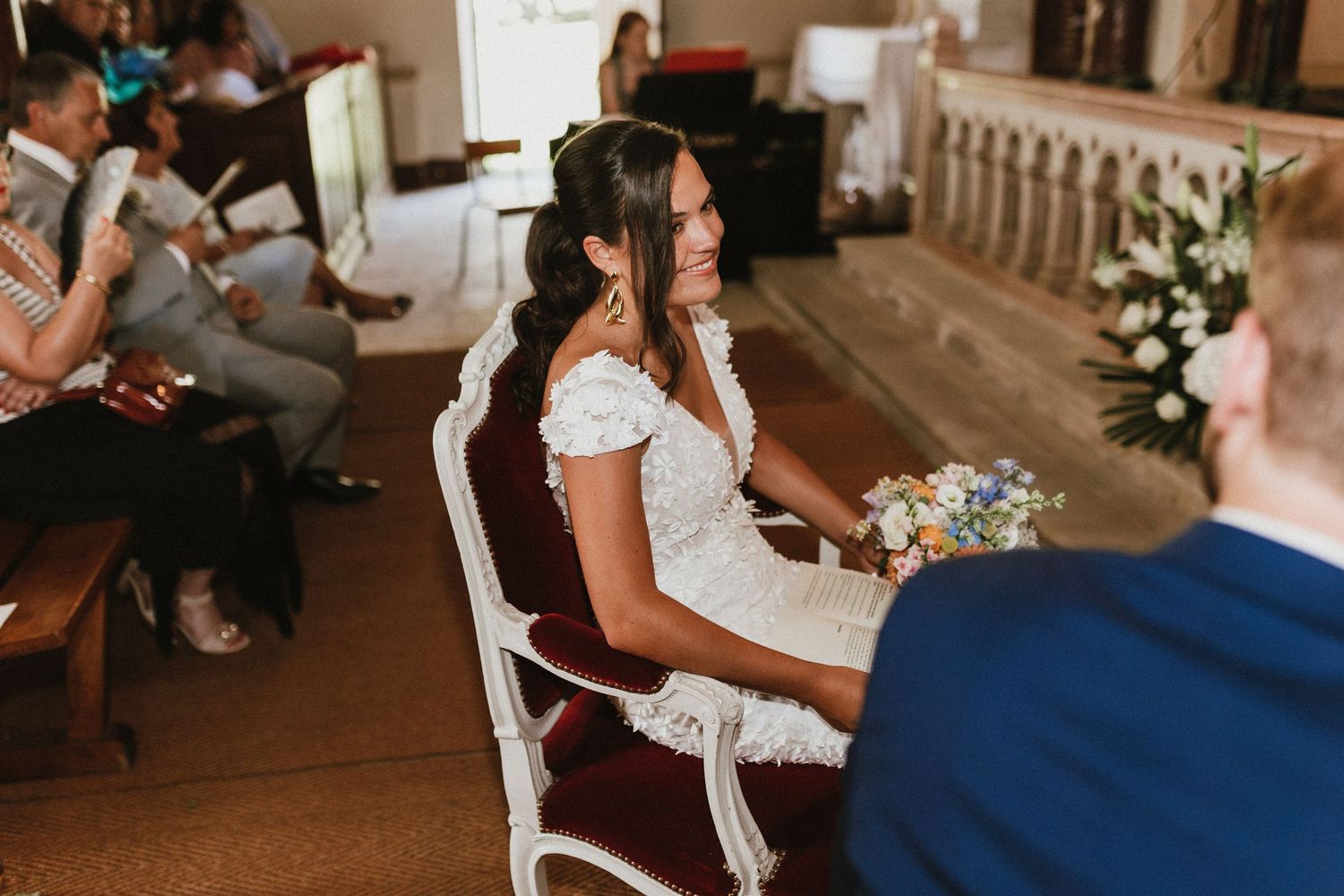 Bride in lace dress sitting during wedding ceremony in church with decorative floral arrangements.
