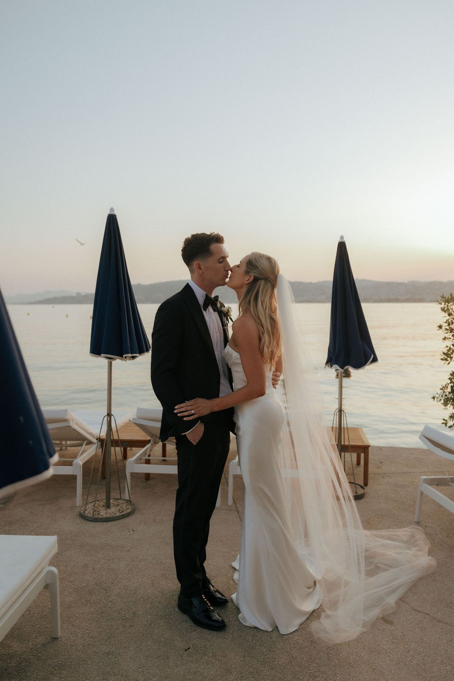 A romantic sunset wedding photo at a seaside venue with navy blue umbrellas and lounge chairs in the background.