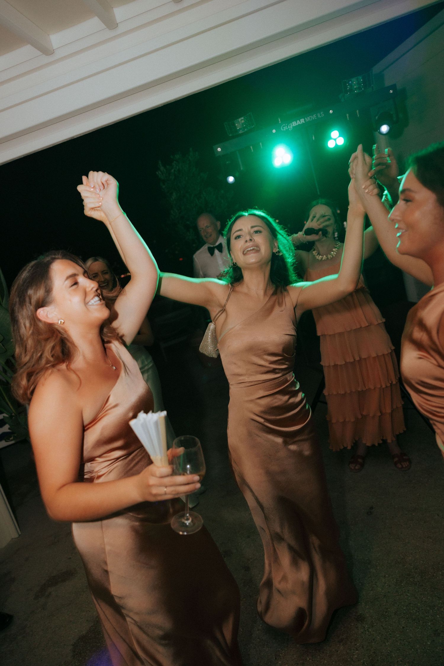 Wedding guests in brown dresses dance energetically under green party lights at the reception.