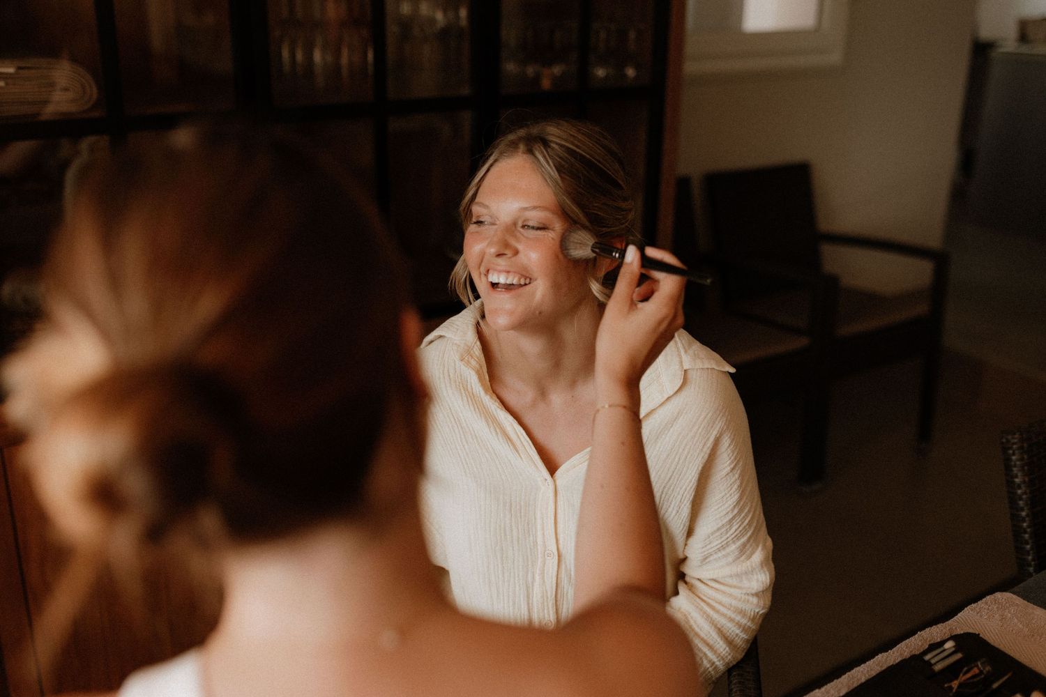 A candid moment shows someone getting professionally styled for a photoshoot, sharing a laugh in warm lighting.