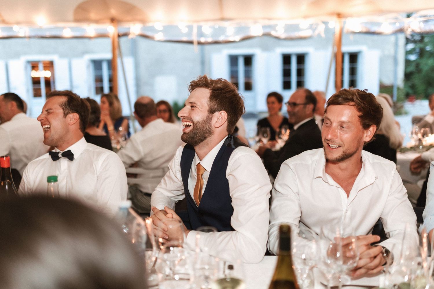 Wedding reception guests enjoying themselves under a white tent with string lights.