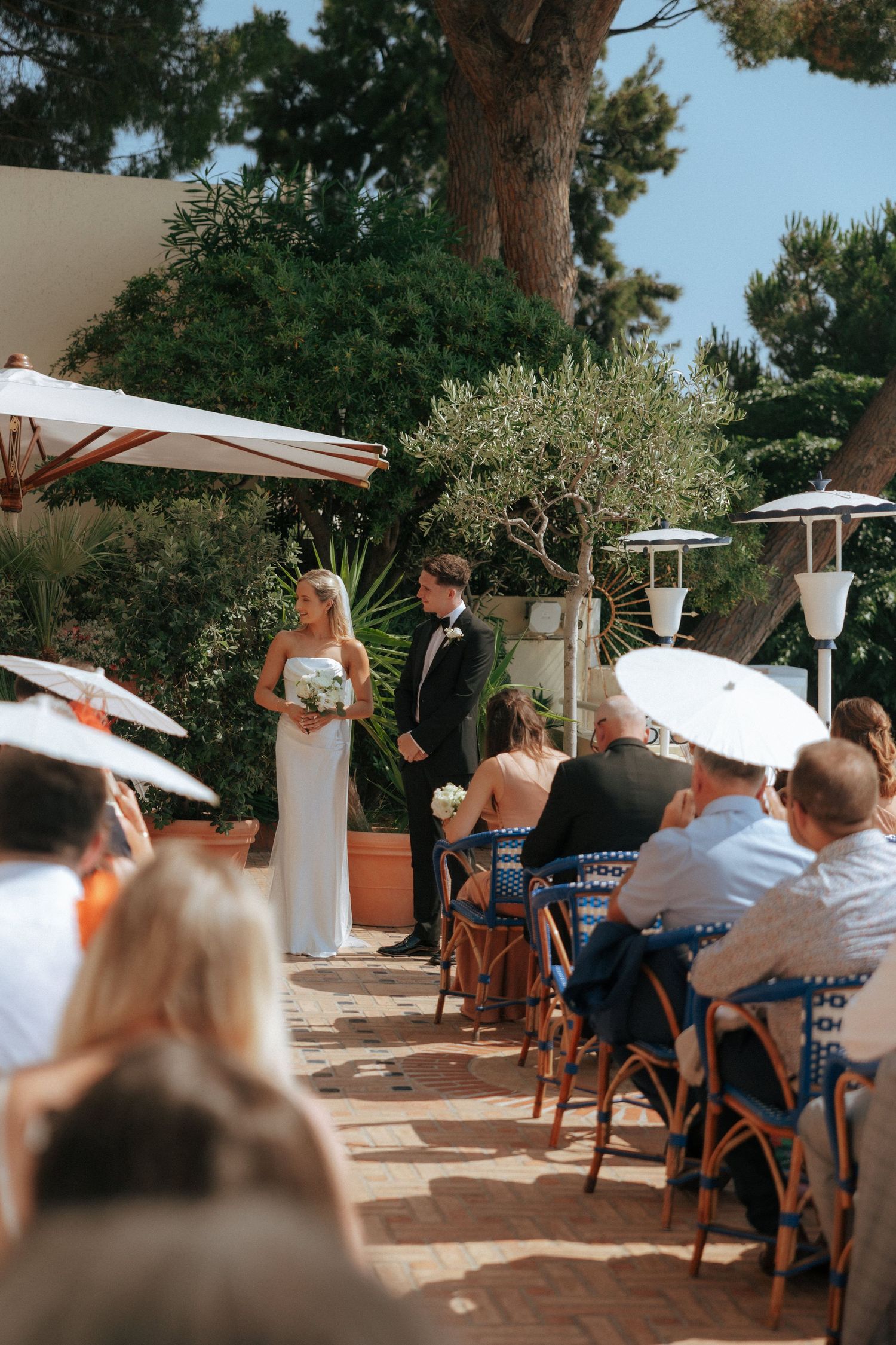 Wedding ceremony guests seated with white parasols under trees at an outdoor venue.