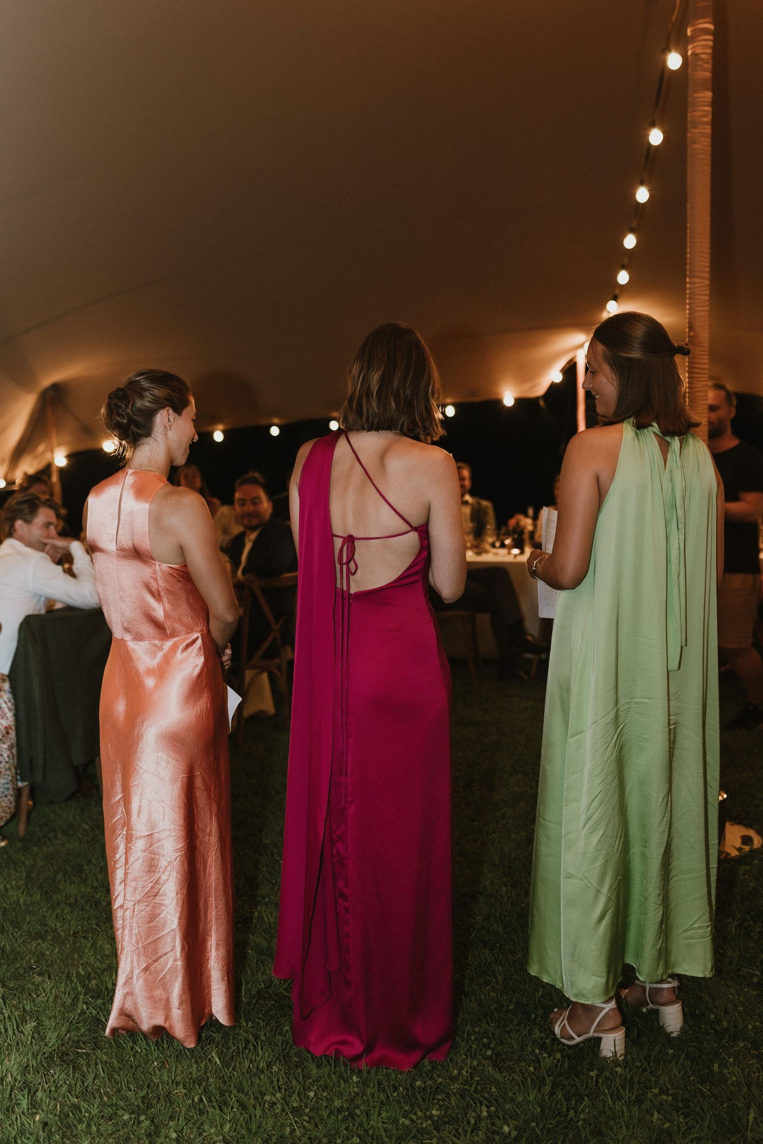 Three women in formal dresses of pink, burgundy and sage green at evening outdoor event.