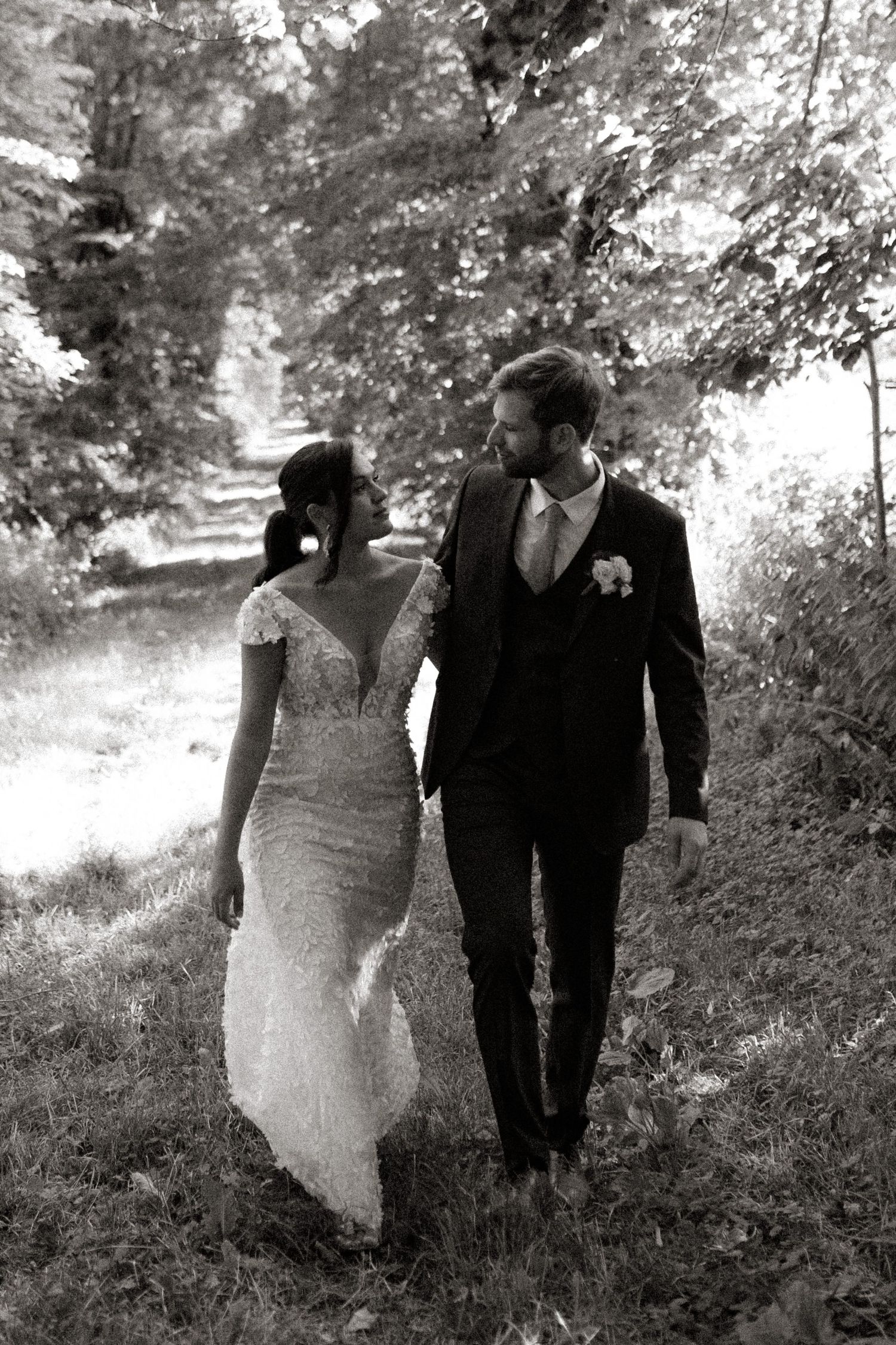 Black and white photo of couple walking along a tree-lined path in wedding attire.