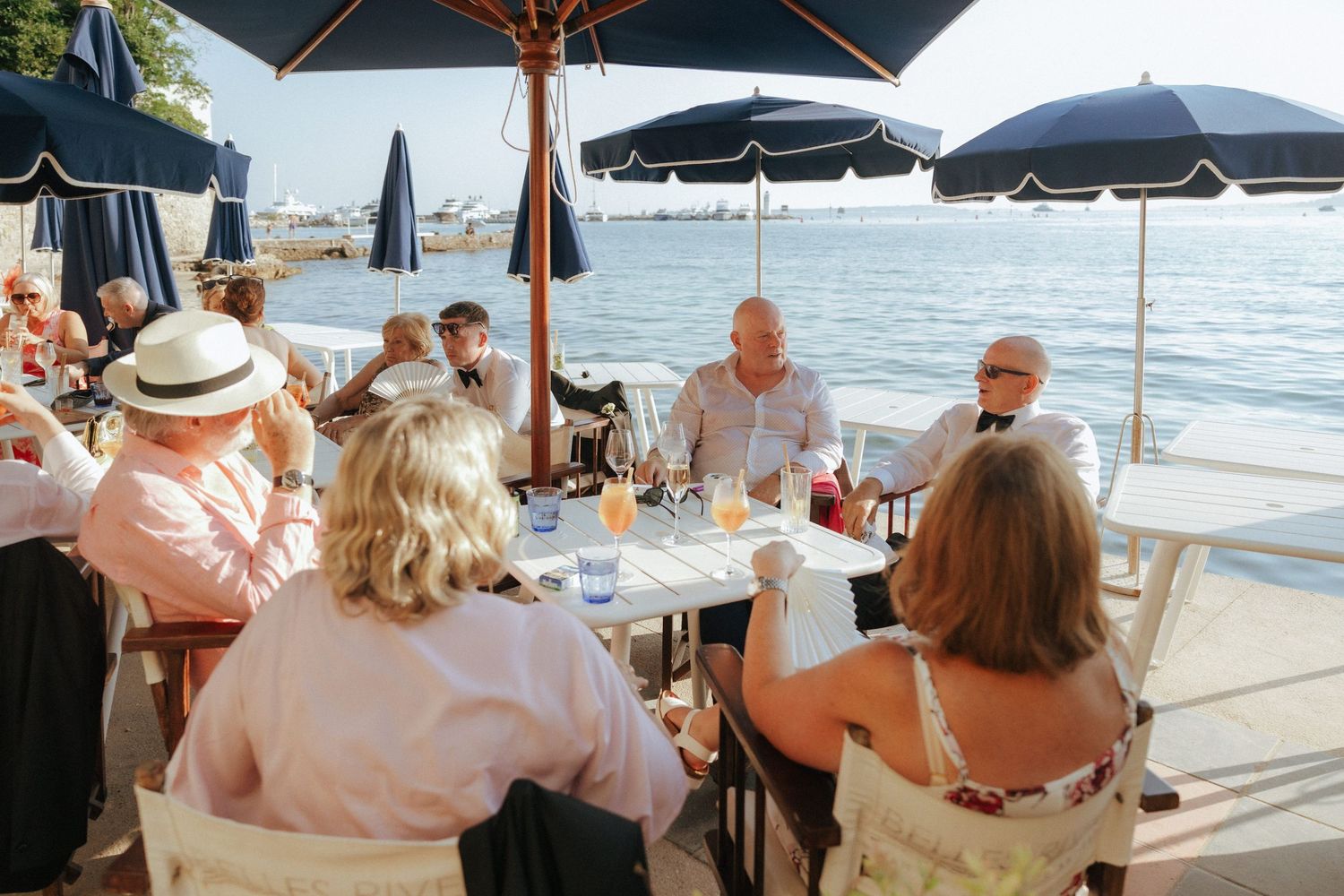 A group of people enjoying lunch at an oceanfront restaurant under blue umbrellas on a sunny afternoon.