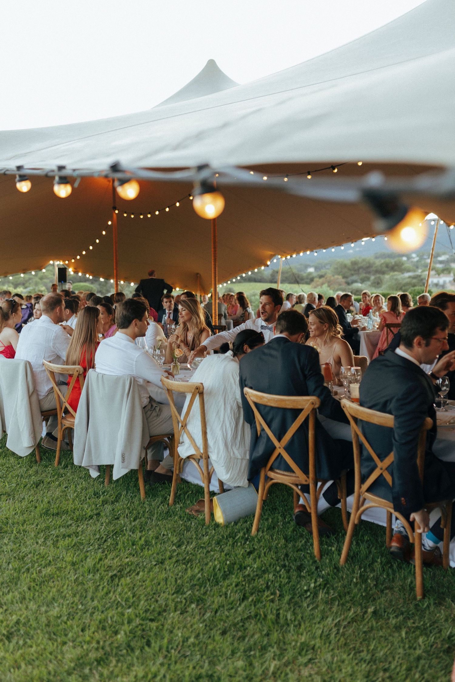 Outdoor wedding reception under white tent with string lights and guests seated at long tables.