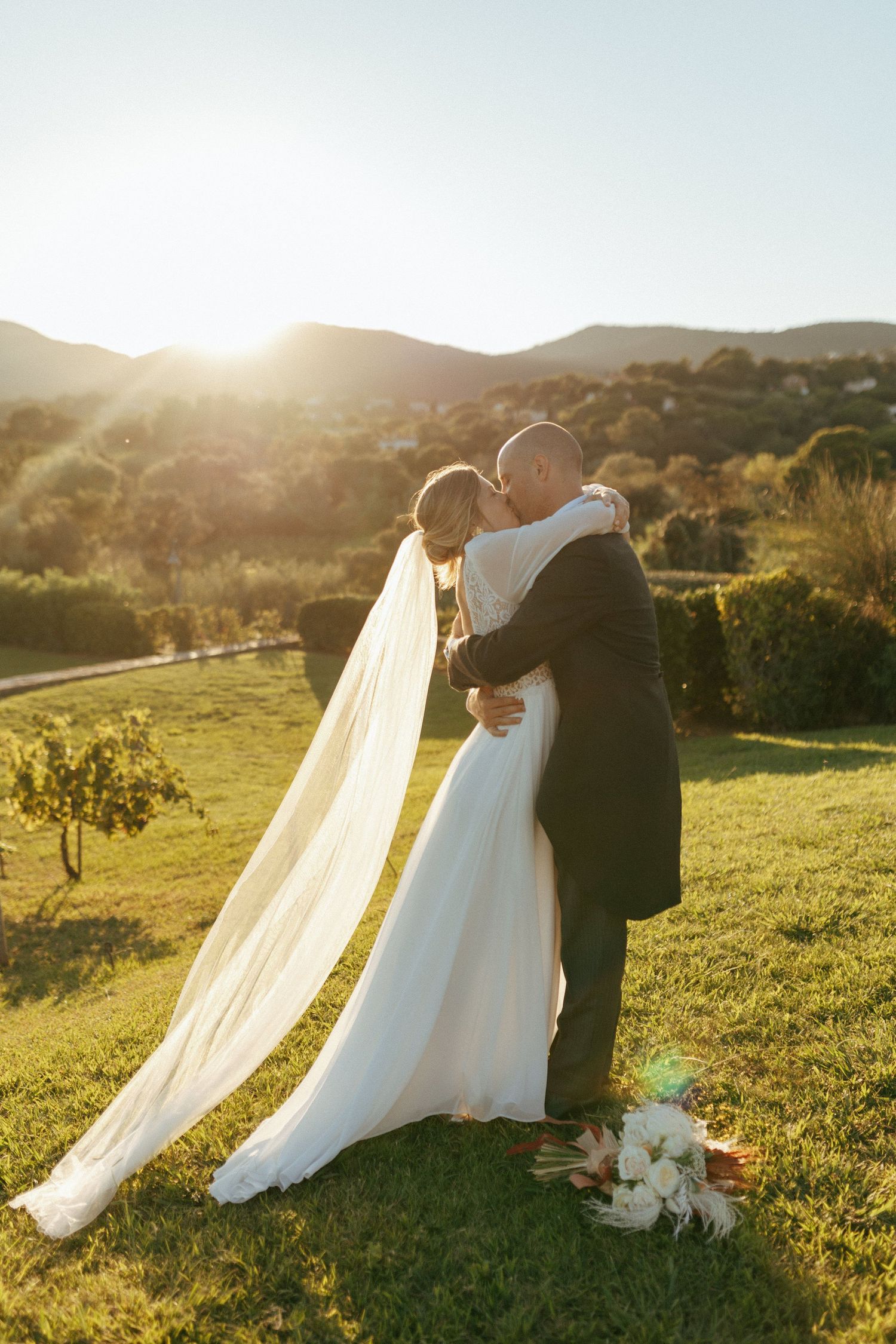 A newlywed couple embraces against a scenic sunset backdrop in a hillside vineyard.