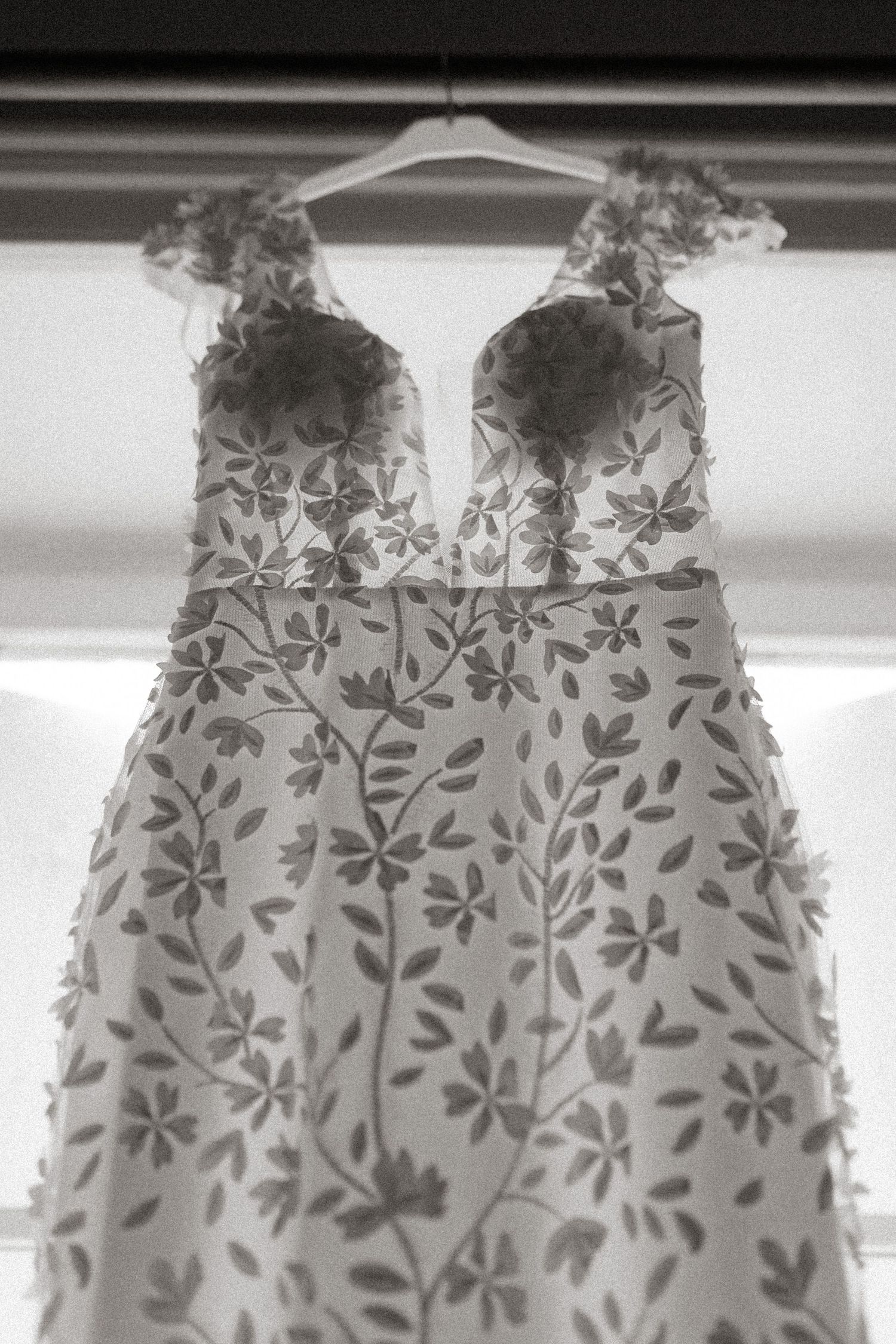 Delicate floral lace wedding dress with plunging neckline hangs against white wall in black and white.