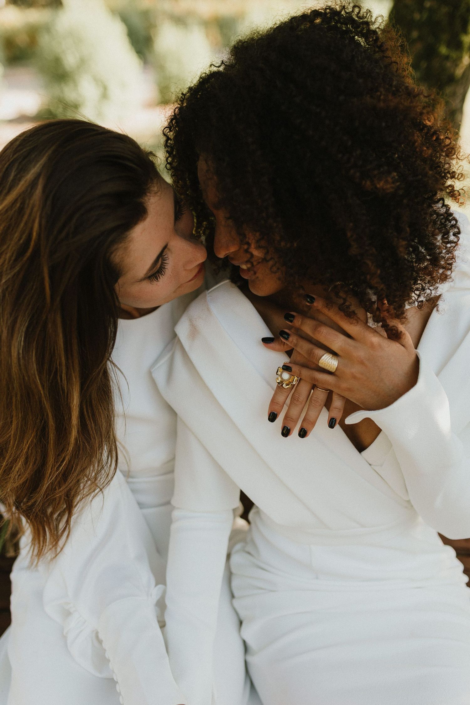 Two people in white outfits share an intimate moment together outdoors, with their hands clasped and heads close.