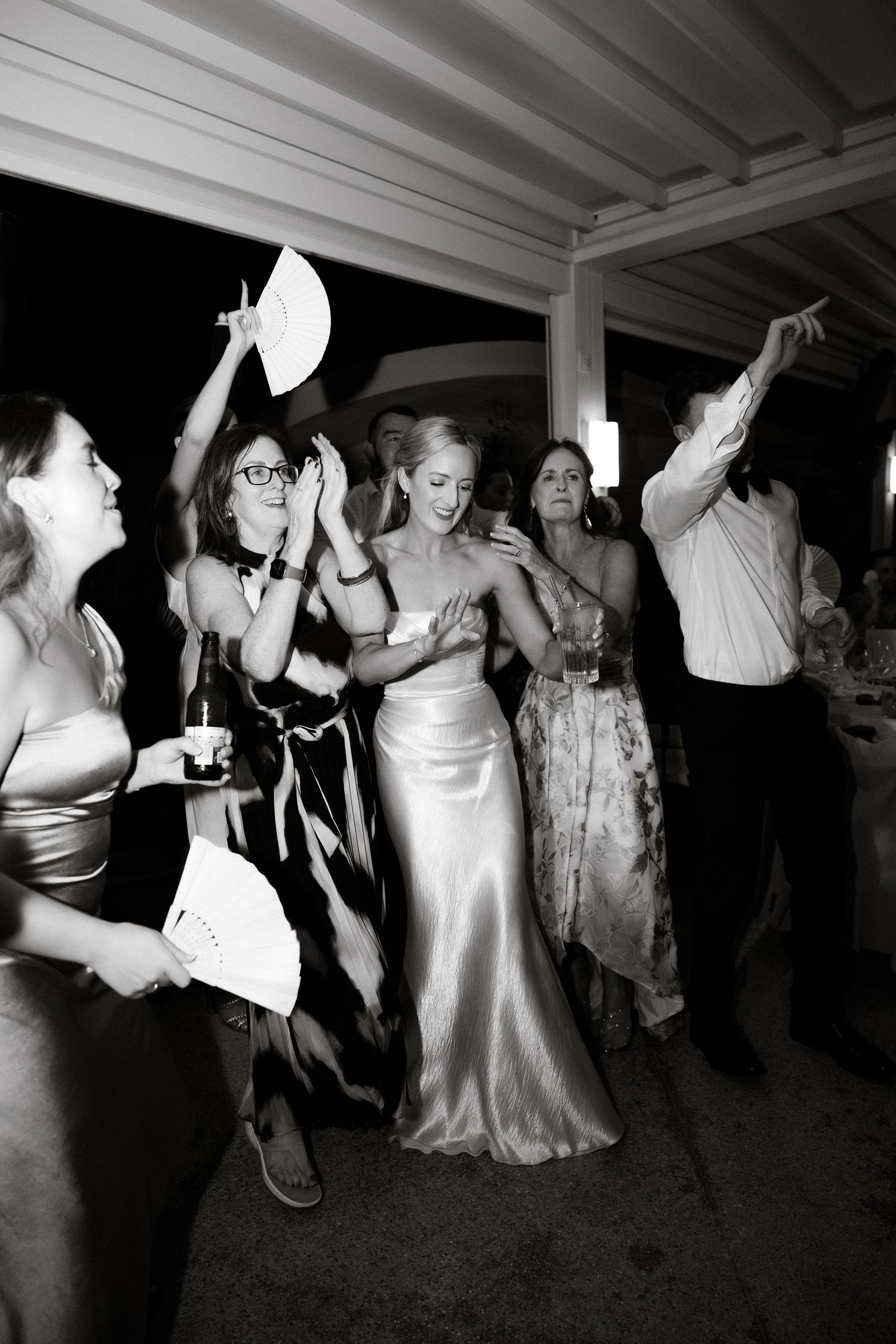 Joyful wedding celebration captured in black and white as guests dance with fans at an evening reception.