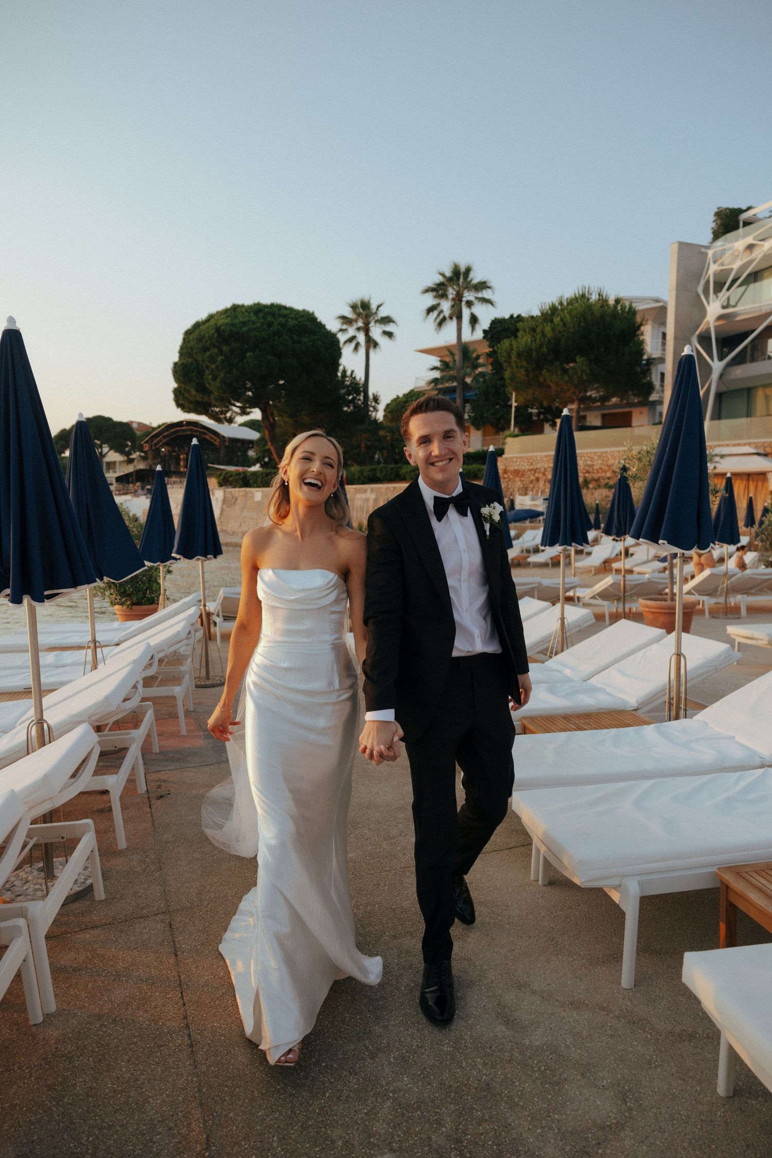 Couple walking between white lounge chairs on a beach club patio during a wedding celebration.