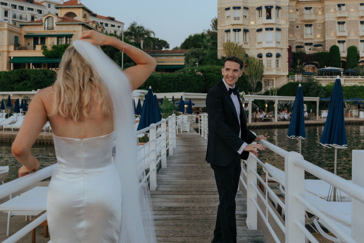 A couple in formal wedding attire stand together on a seaside pier with coastal buildings in the background.