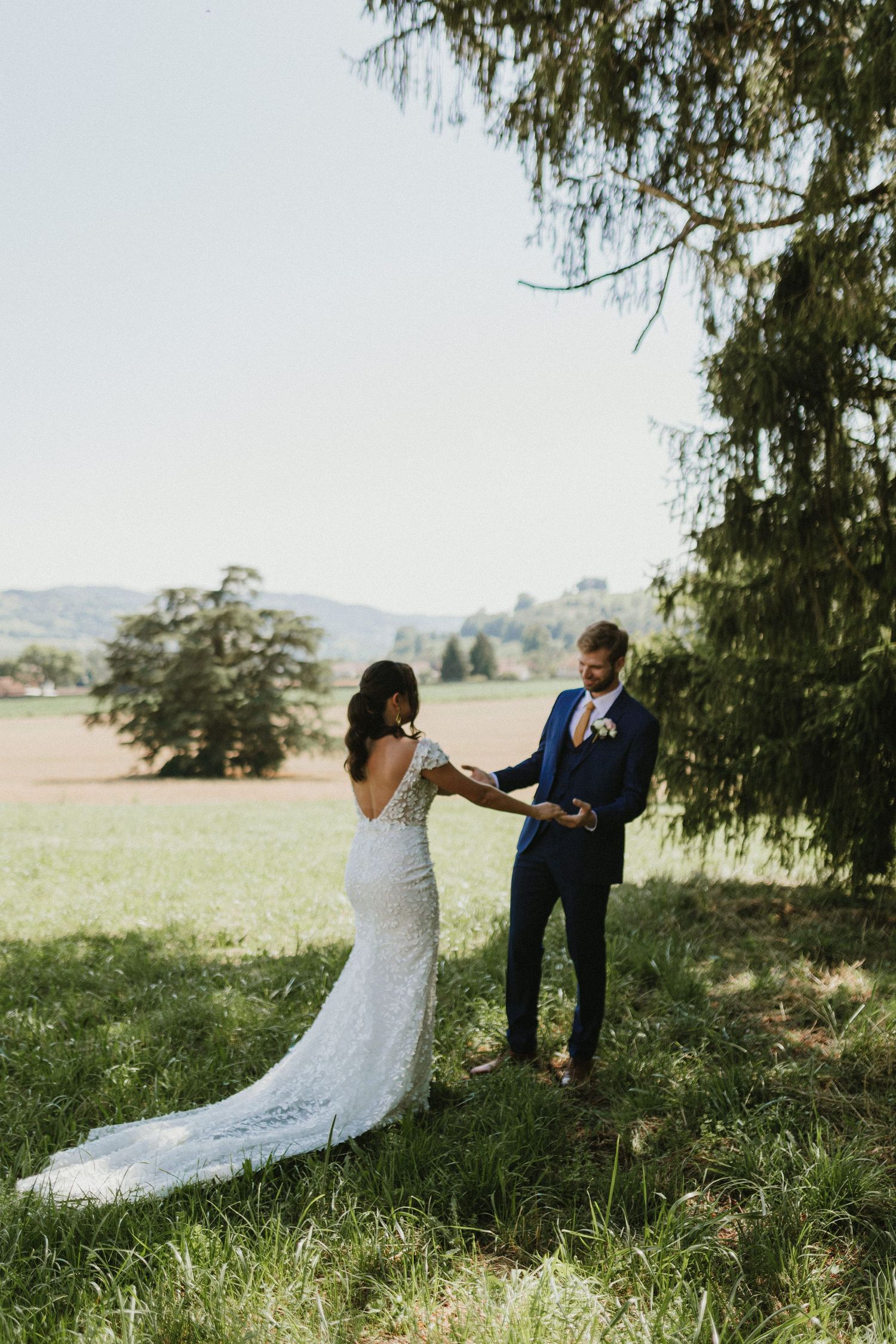 A couple shares a romantic moment under a tree in their wedding attire on a scenic hillside.
