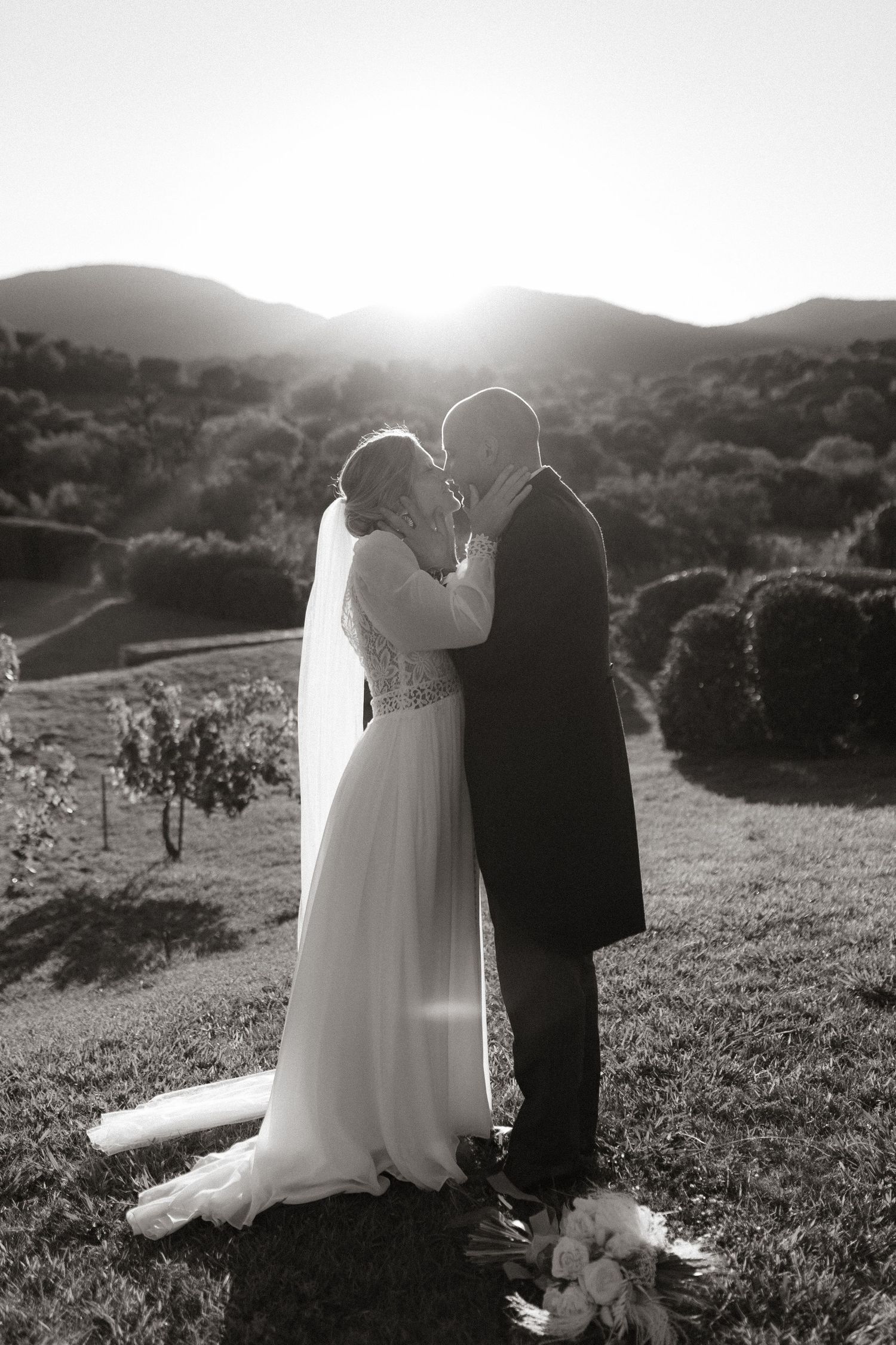 A romantic black and white silhouette of newlyweds sharing a moment at sunset on a hilltop with mountains in background.