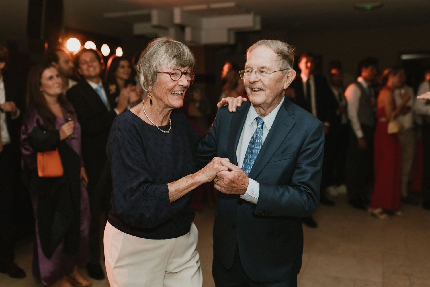 Two elderly people dance together at a dimly lit formal event while other attendees mingle in the background.