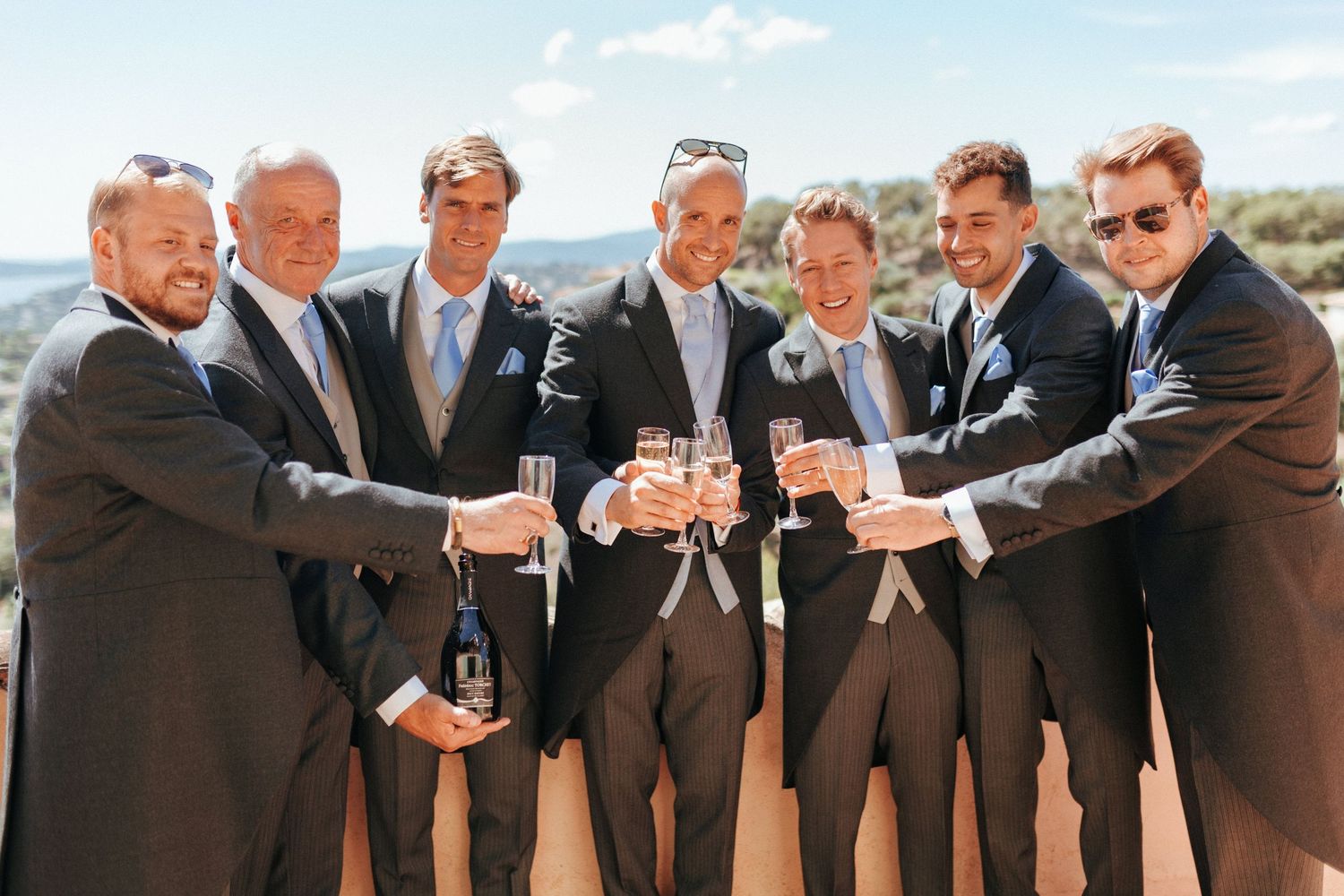 Groomsmen in dark suits toasting with champagne glasses against a mountain backdrop.