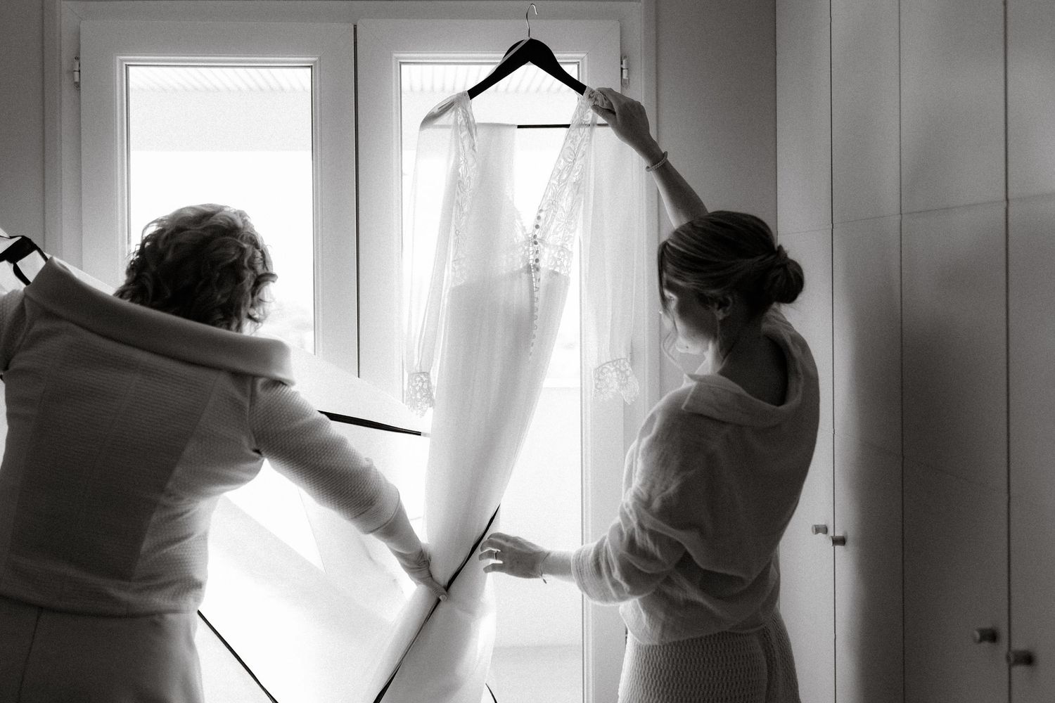 Two people prepare a white wedding dress hanging near a bright window in a soft black and white photograph.