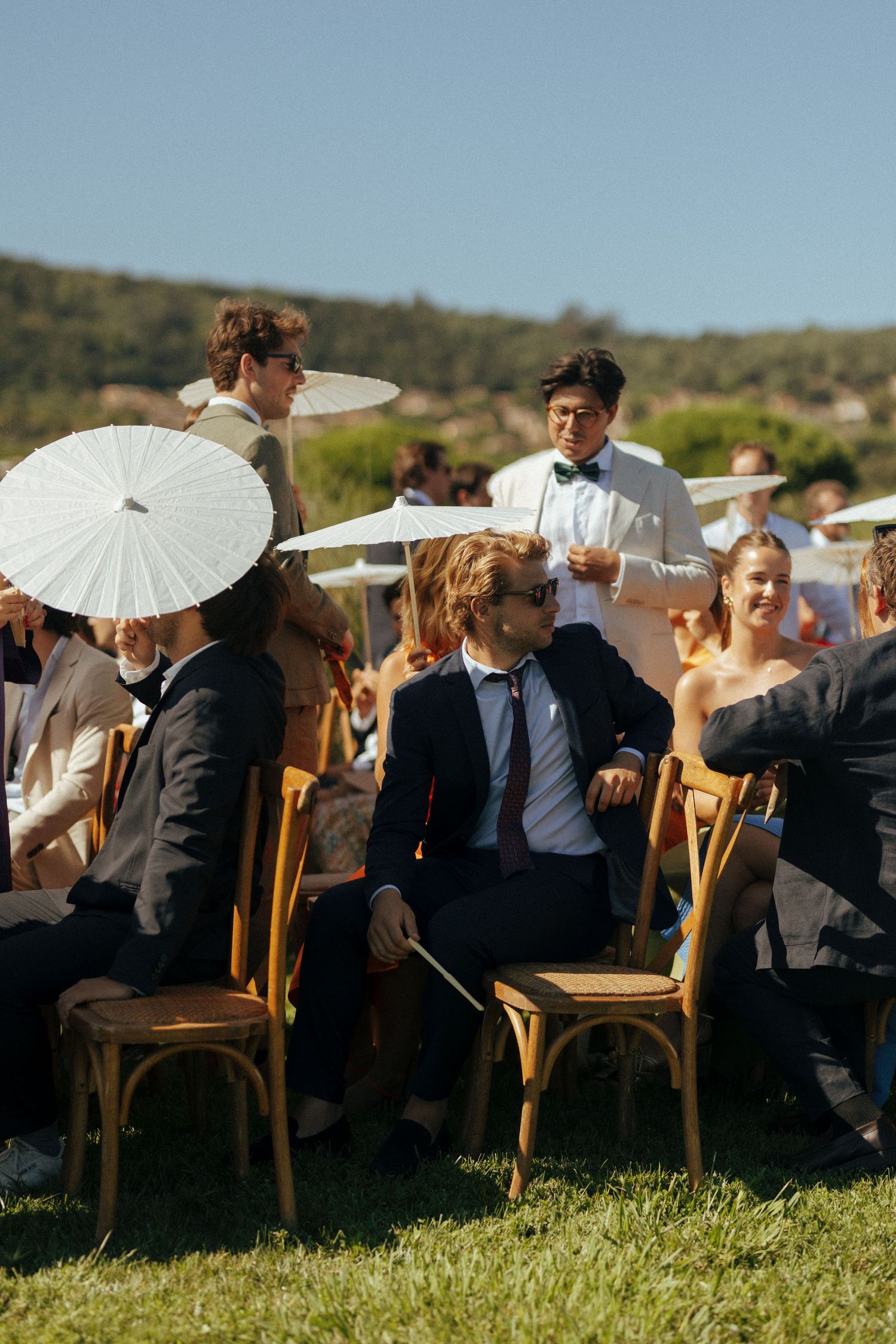 Wedding guests sit under parasols at an outdoor ceremony in a countryside setting.