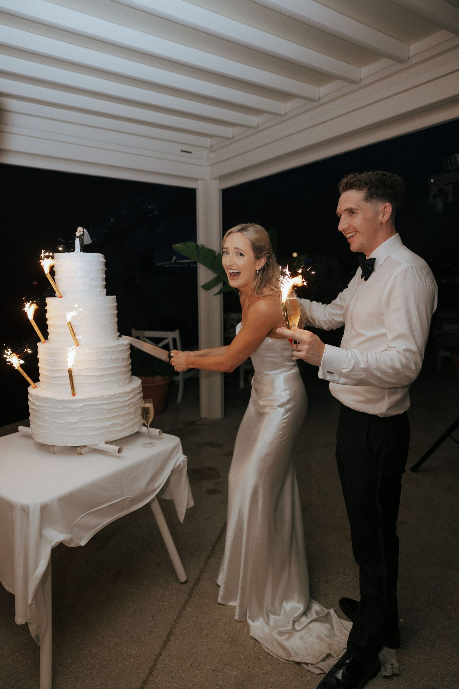 A wedding couple light sparklers on their elegant white tiered cake at night.