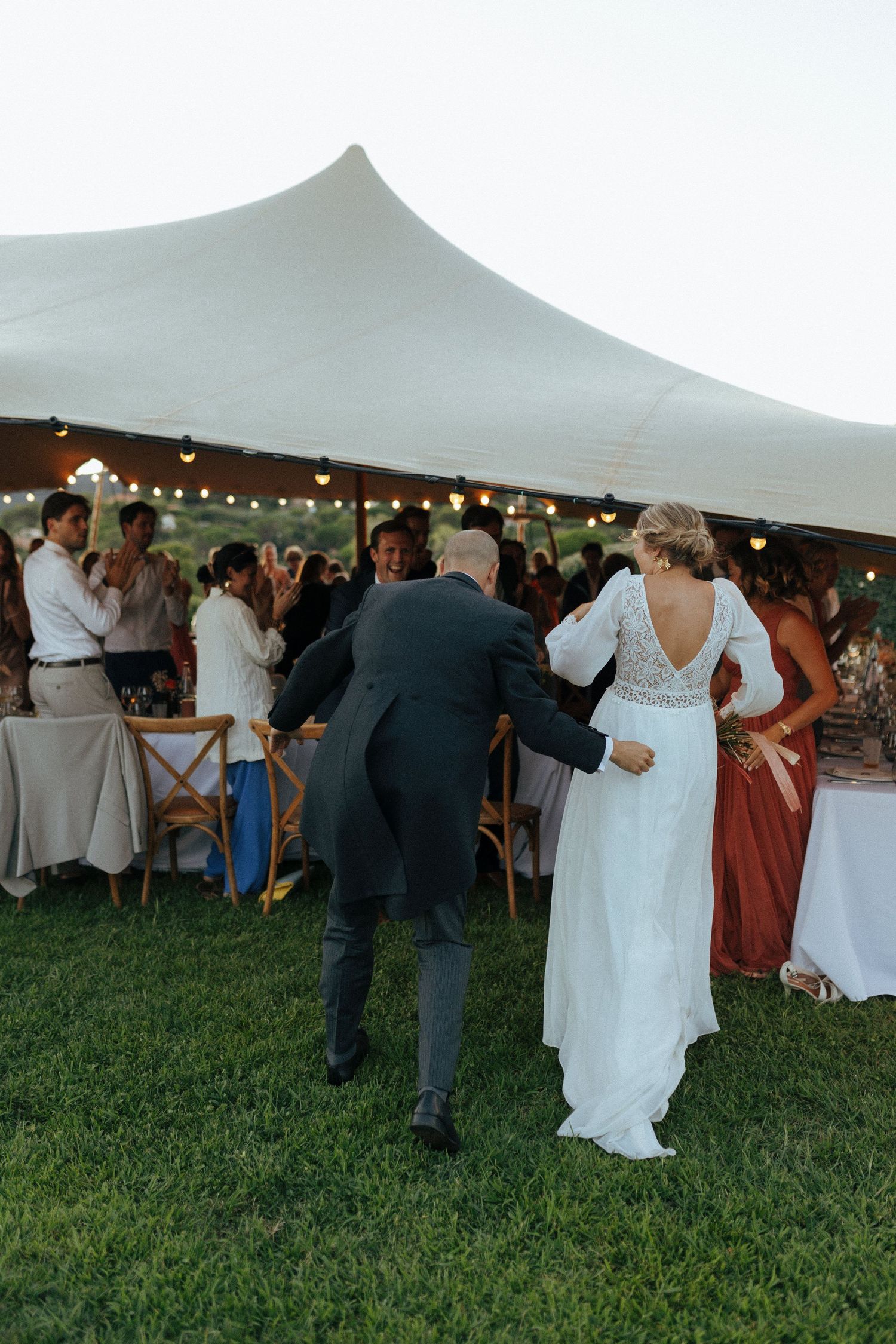 Wedding reception under white tent with string lights and guests seated at tables on grass.