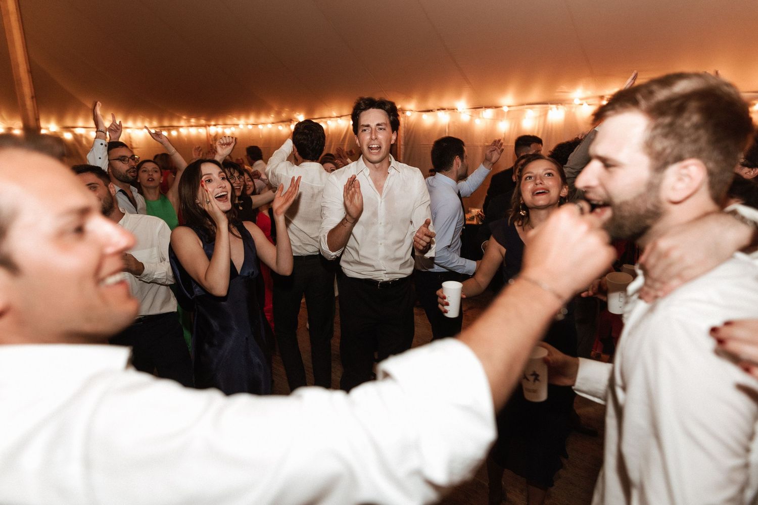 Energetic wedding guests dancing and celebrating under a reception tent with ambient lighting.
