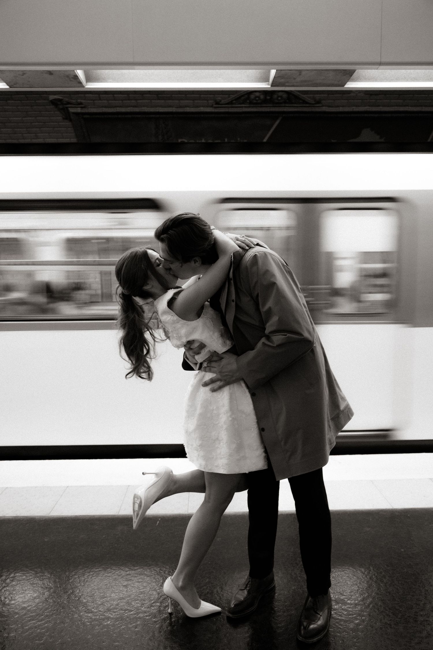 A romantic couple shares a passionate kiss at a subway platform as a blurred train rushes past in black and white.