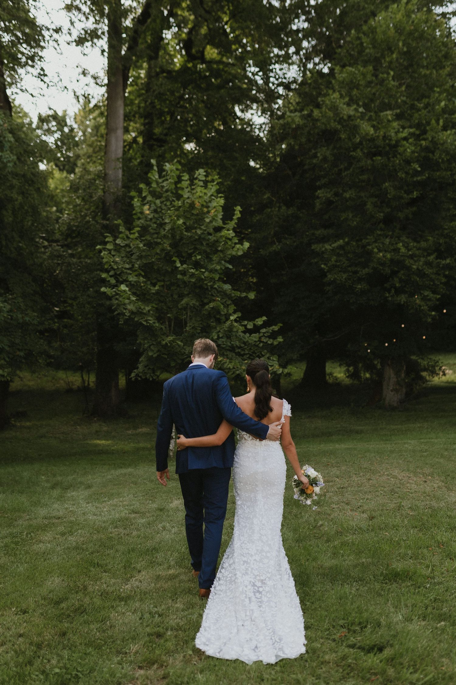 Newlywed couple walking away holding each other on grass with forest backdrop.