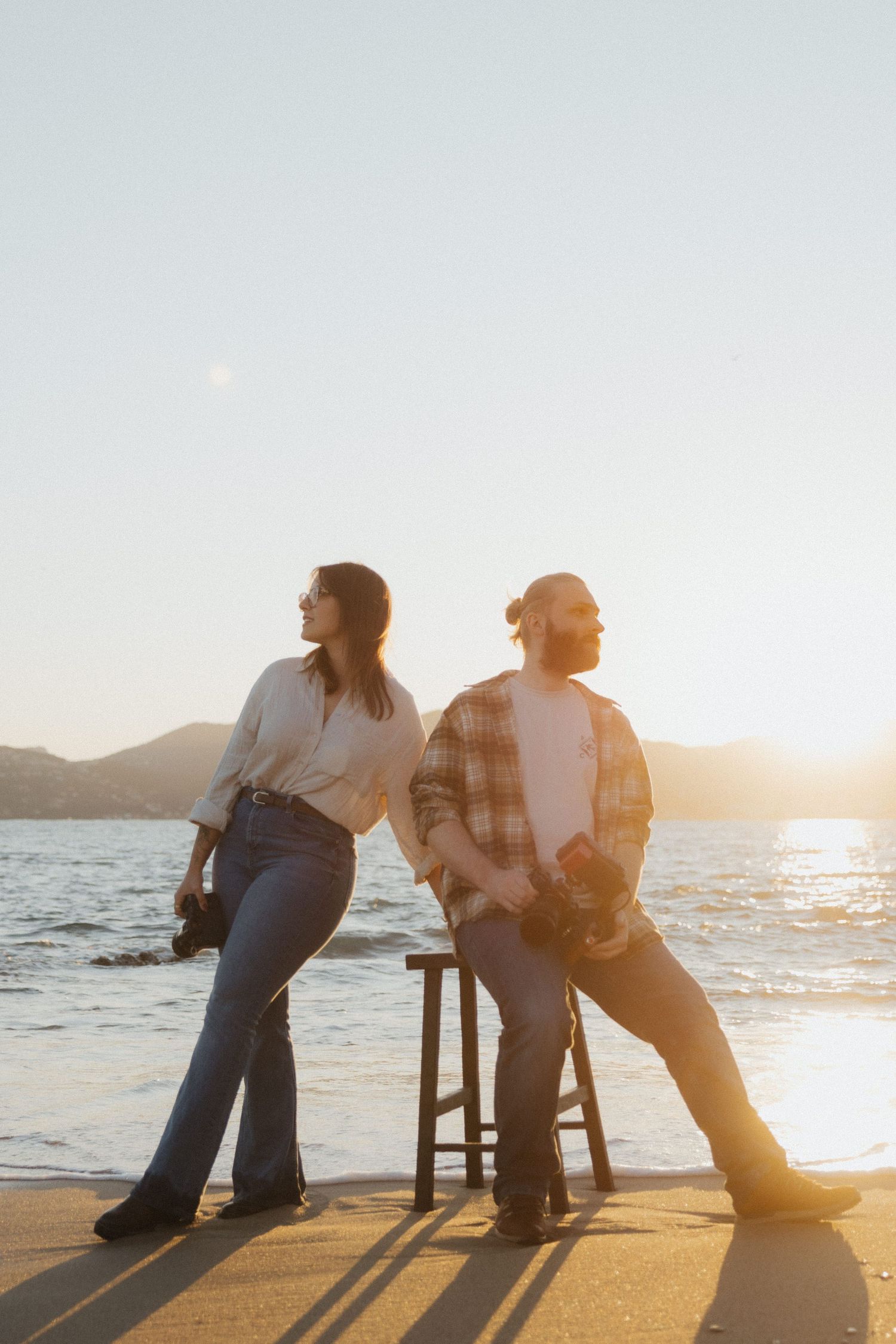 Two silhouettes lean against a barstool at sunset by the ocean.