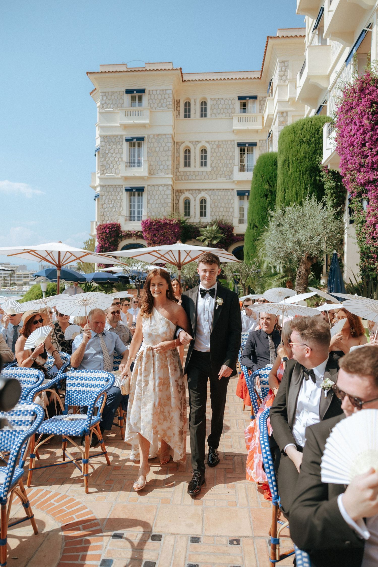 Wedding celebration on a Mediterranean terrace with blue and white checkered chairs and umbrellas against stone villa backdrop.