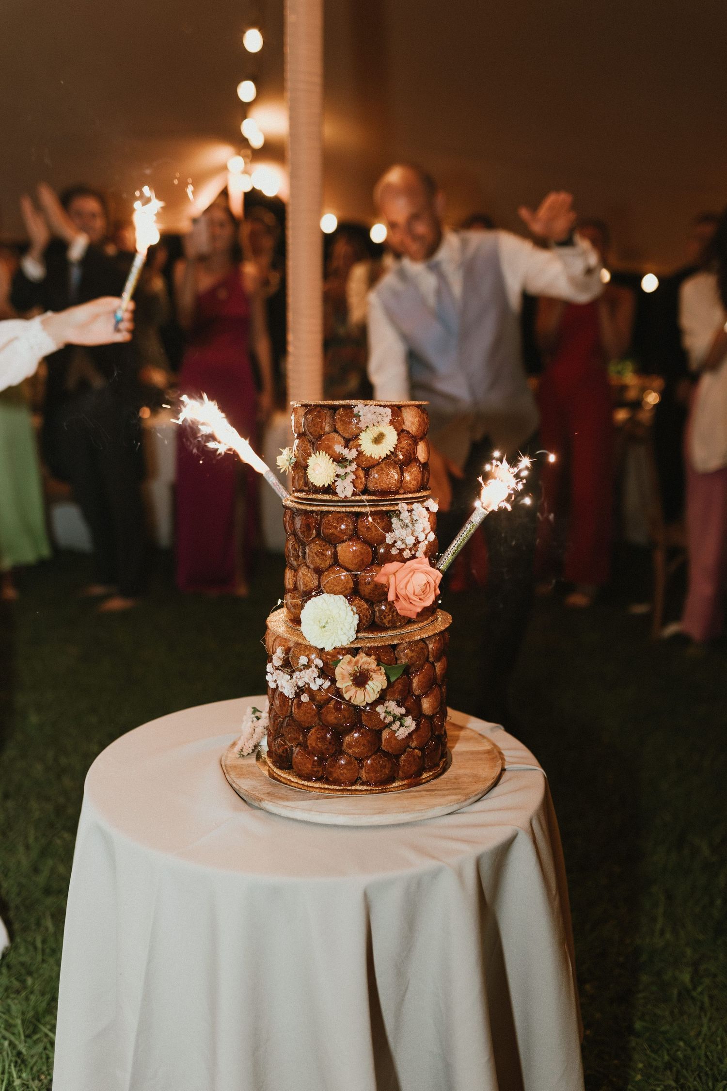 Three-tiered chocolate wedding cake decorated with macarons and sparklers on white tablecloth at evening reception.
