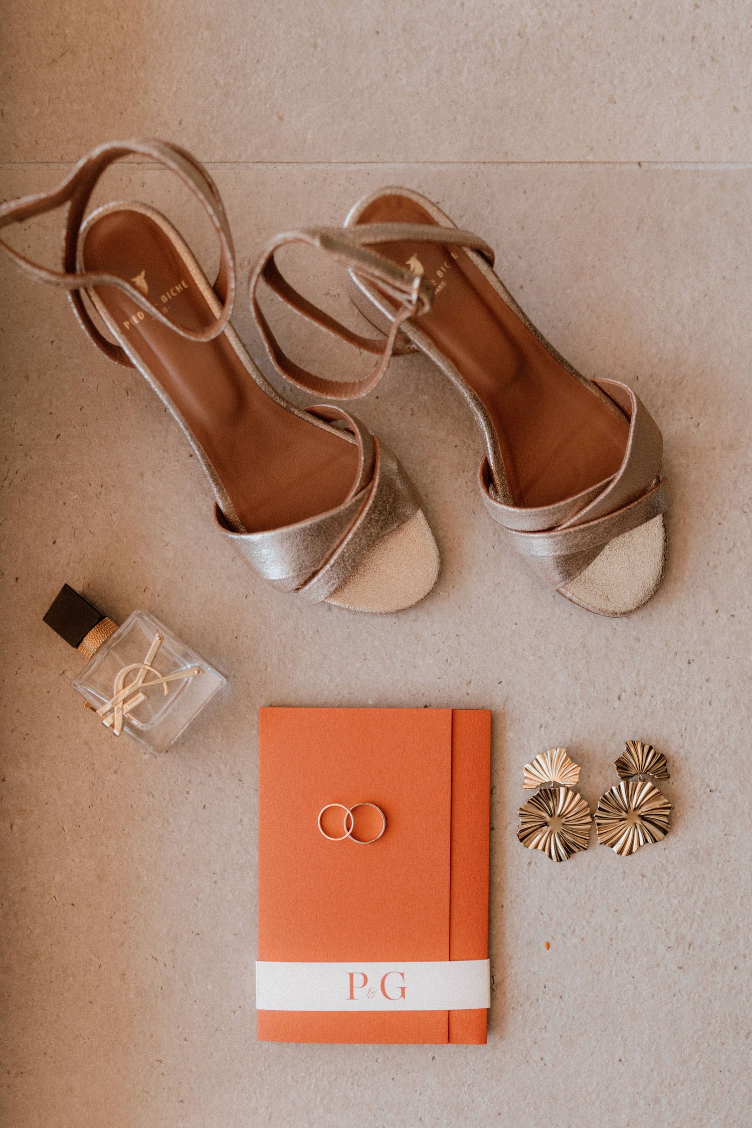 Wedding accessories including silver heels, perfume bottle, orange monogrammed book and gold sunburst earrings on beige surface.