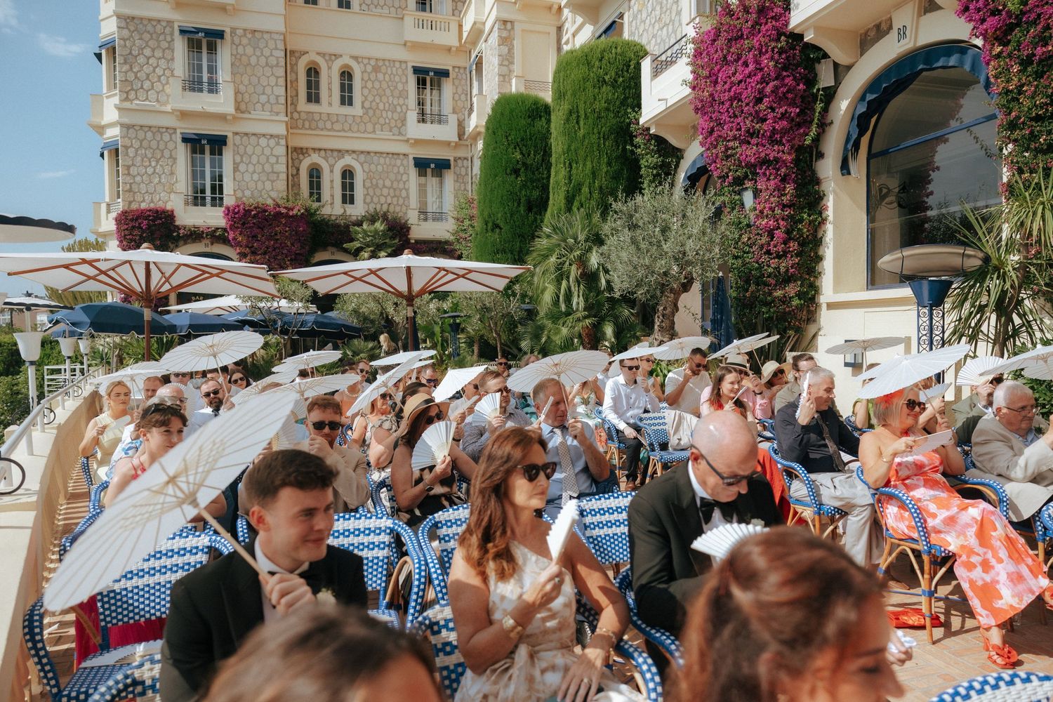 A crowd of outdoor wedding guests sitting in rows at a Mediterranean venue with vibrant bougainvillea and cypress trees.
