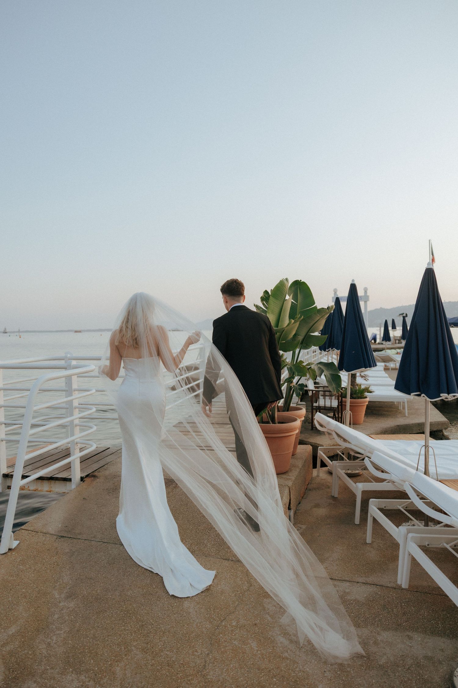A wedding couple strolls along a seaside boardwalk with white chairs and blue umbrellas in the background.