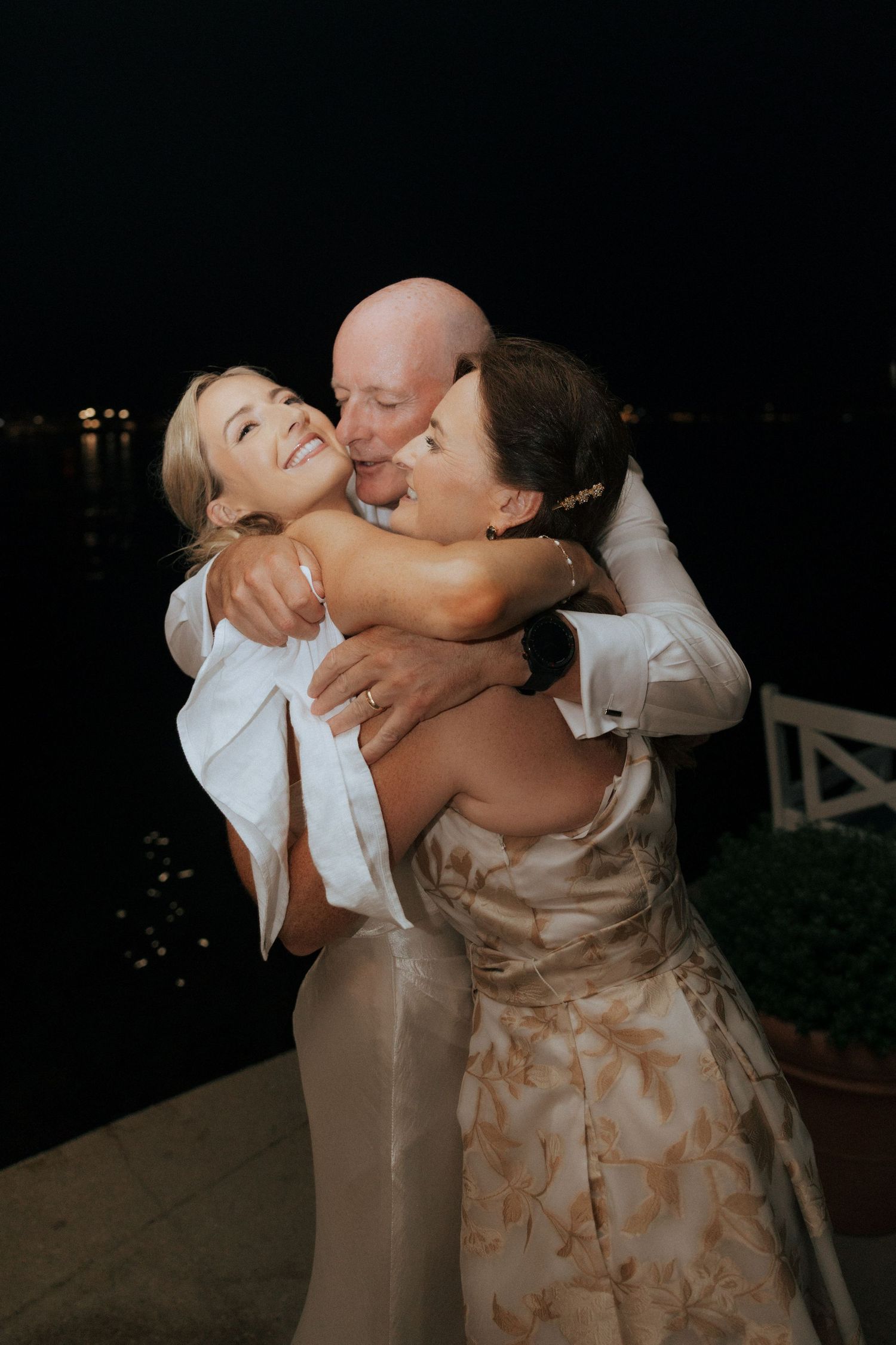 Three people share an emotional embrace during an evening wedding celebration.